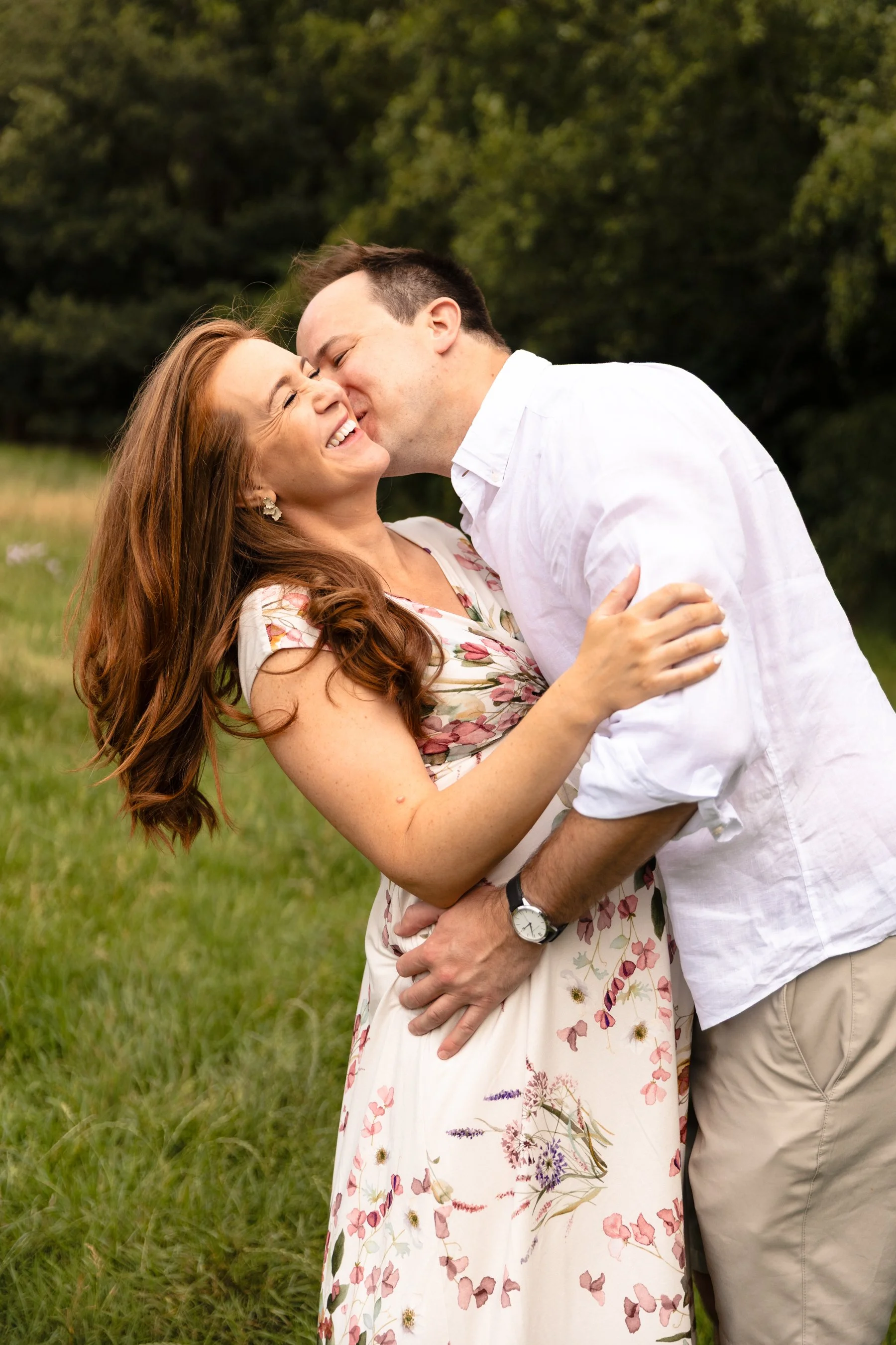 A laughing pregnant couple during an outdoor photoshoot in Wandsworth, as the husband playfully dips his wife back and kisses her neck.