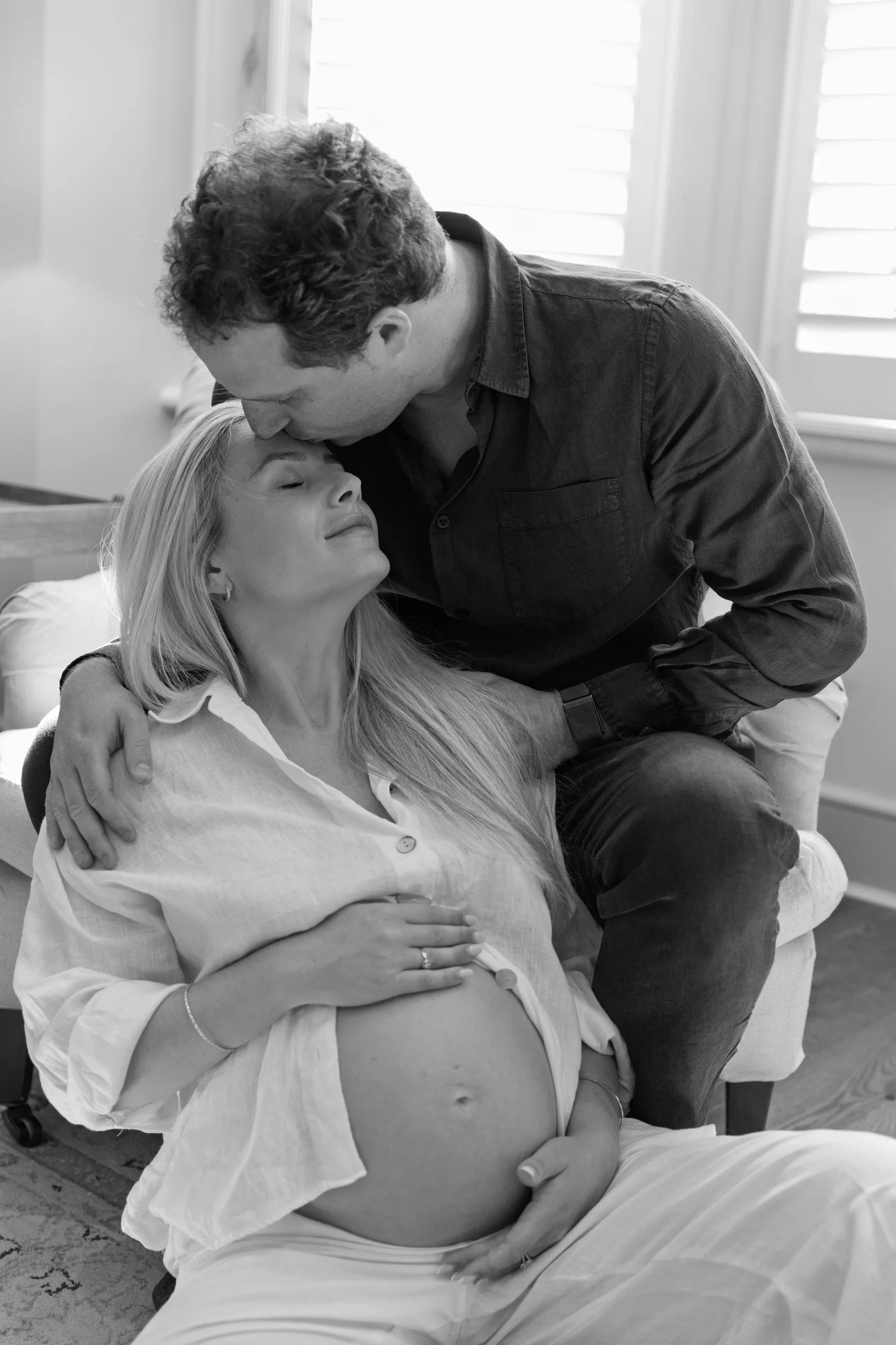 A pregnant woman sitting on the floor between her husband's legs as he sits in an armchair, cradling her bump and looking up as he kisses her forehead during a Wandsworth photoshoot.
