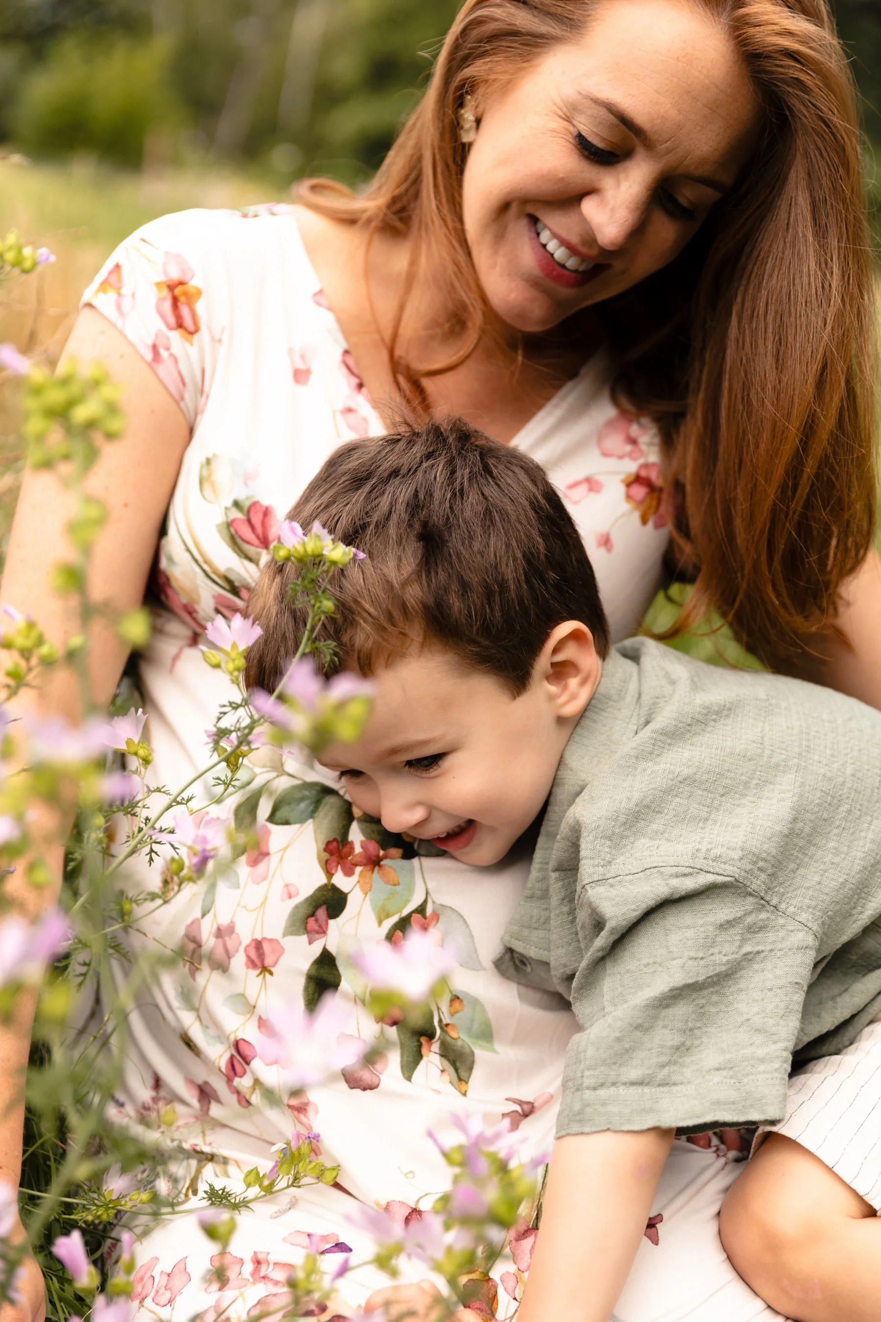 A pregnant mother sitting on the ground looking at her two-year-old son leaning on her bump, with wildflowers blurred in the foreground during an Earlsfield photoshoot