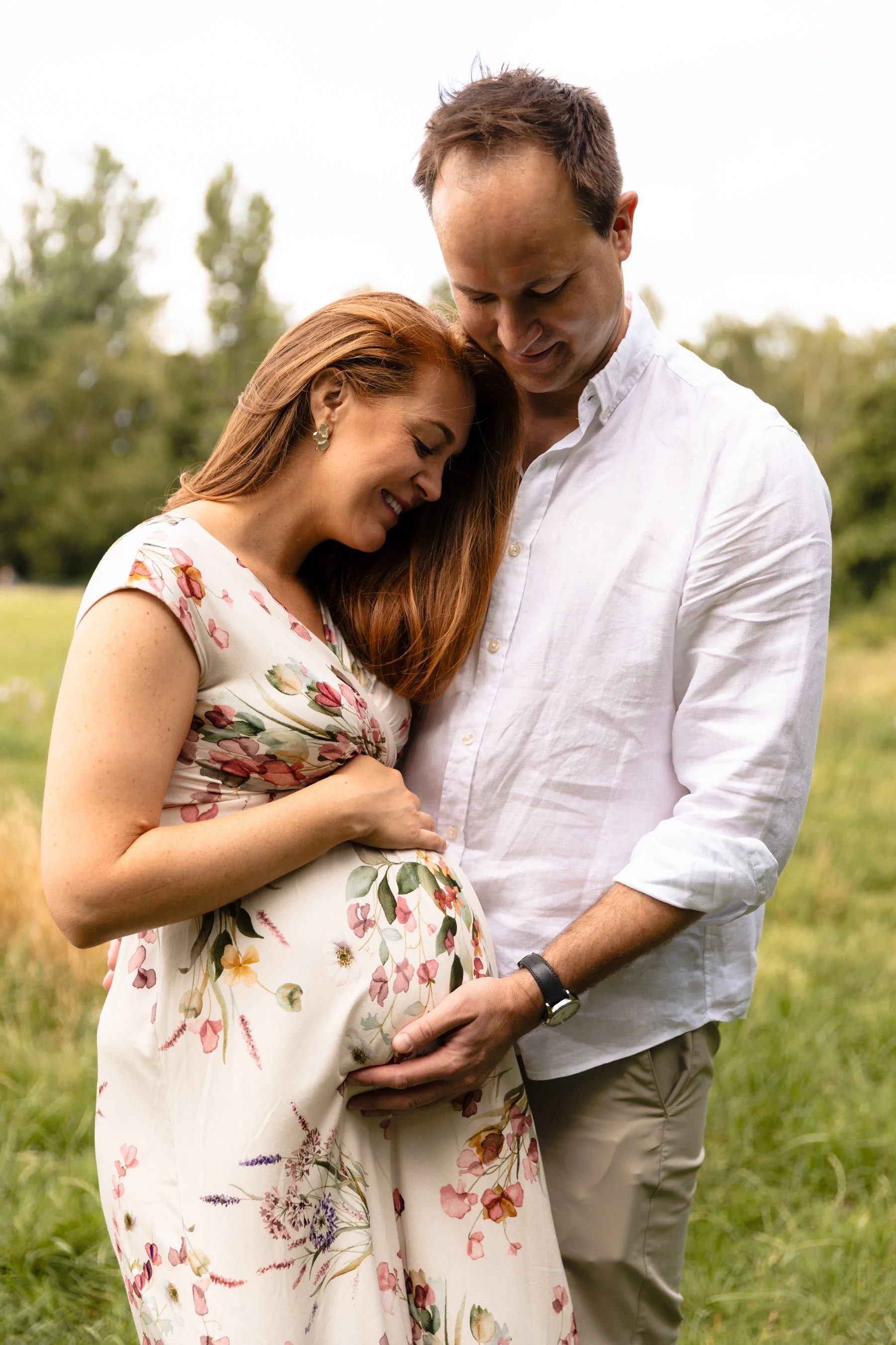 A pregnant couple hold each other close while he cradles the bottom of her bump and she the top at an outdoor maternity photoshoot in Earlsfield