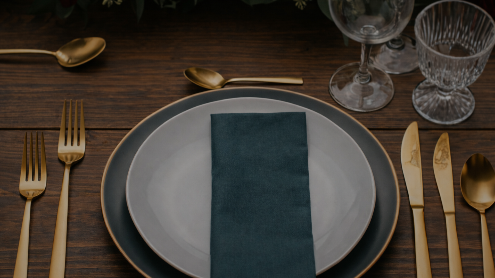 Elegant table setting with gold flatware, a dark blue napkin on white and dark blue plates, and glassware on a wooden table.