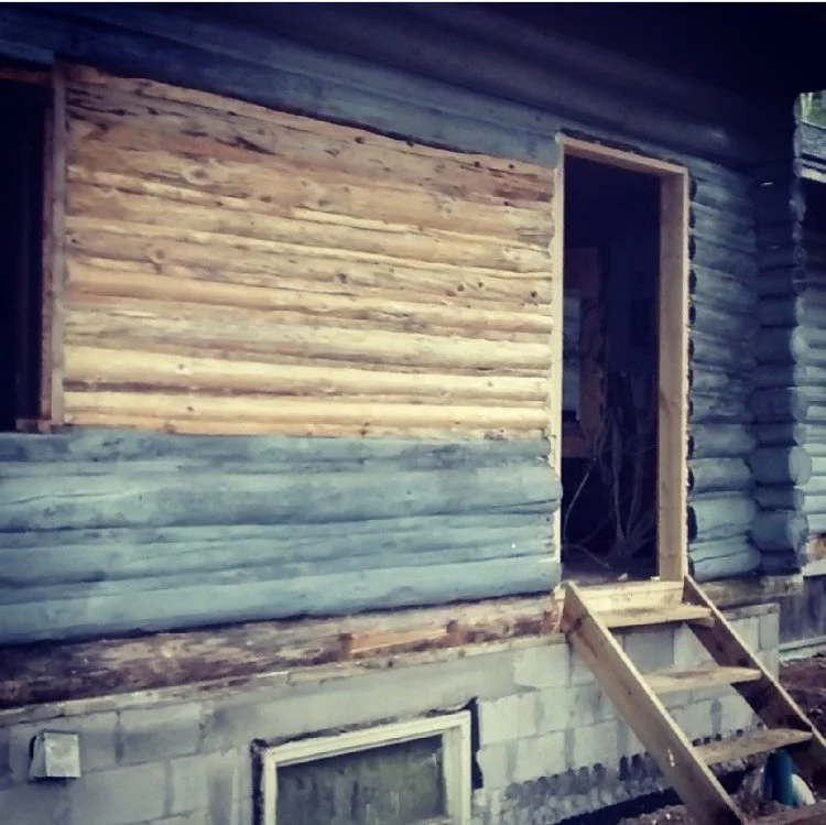A house under construction with partially installed wooden log siding on the exterior walls and a door opening with stairs leading up to it, showing a mix of log siding and concrete blocks.