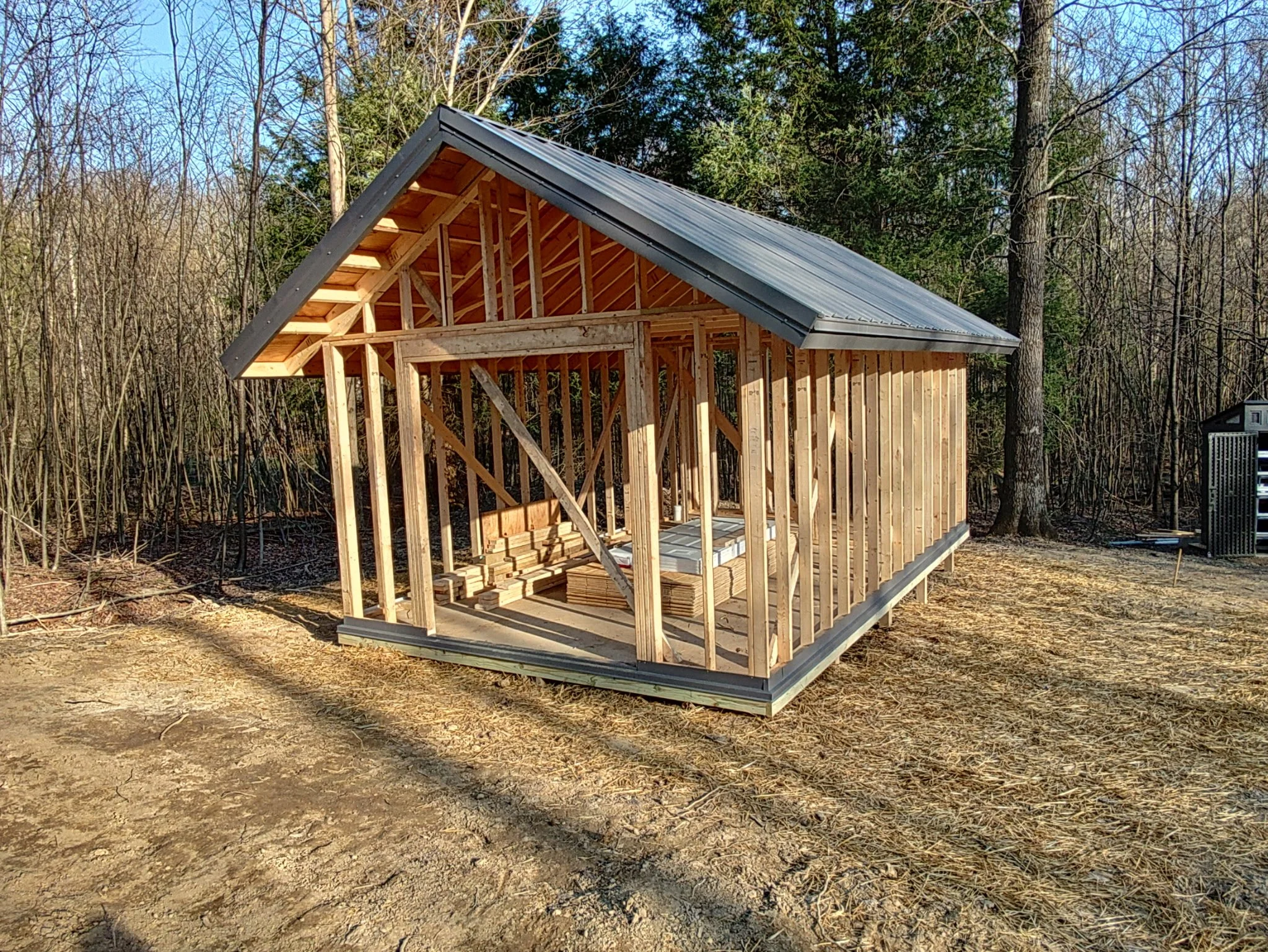 Wooden frame of a shed under construction in a wooded area, with trees in the background and construction materials inside.
