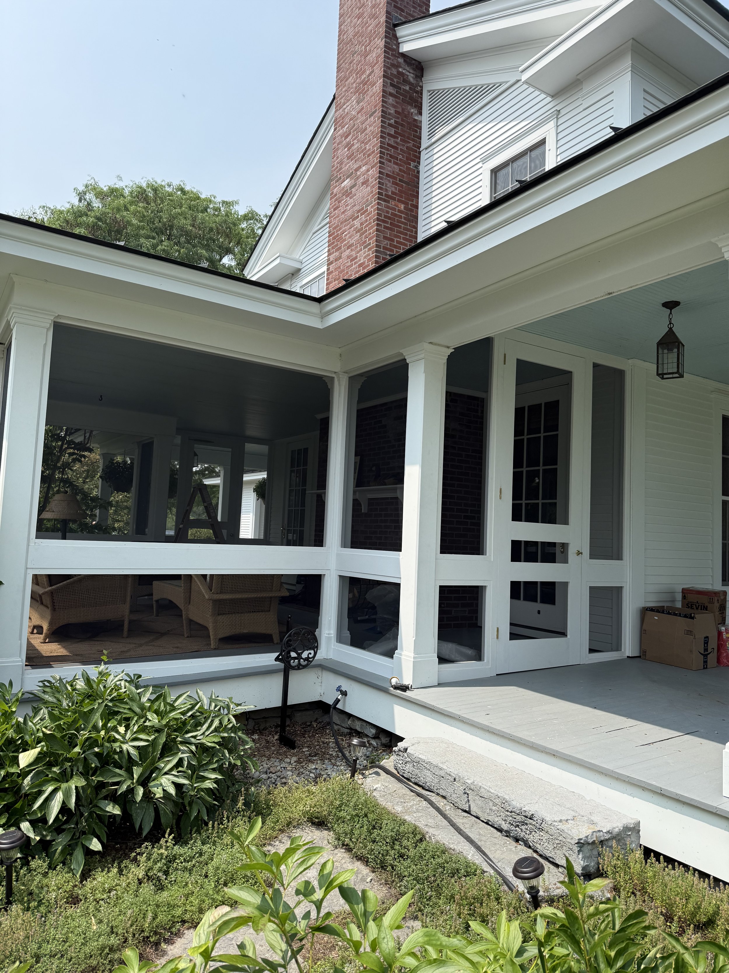 Photo of a house porch with white framing, screened enclosure, patio furniture visible inside, and some boxes on the porch floor. Green shrubs and plants in the foreground.