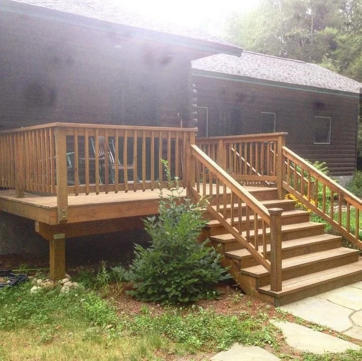 New wooden deck with stairs attached to a dark-colored house, surrounded by green plants and grass.