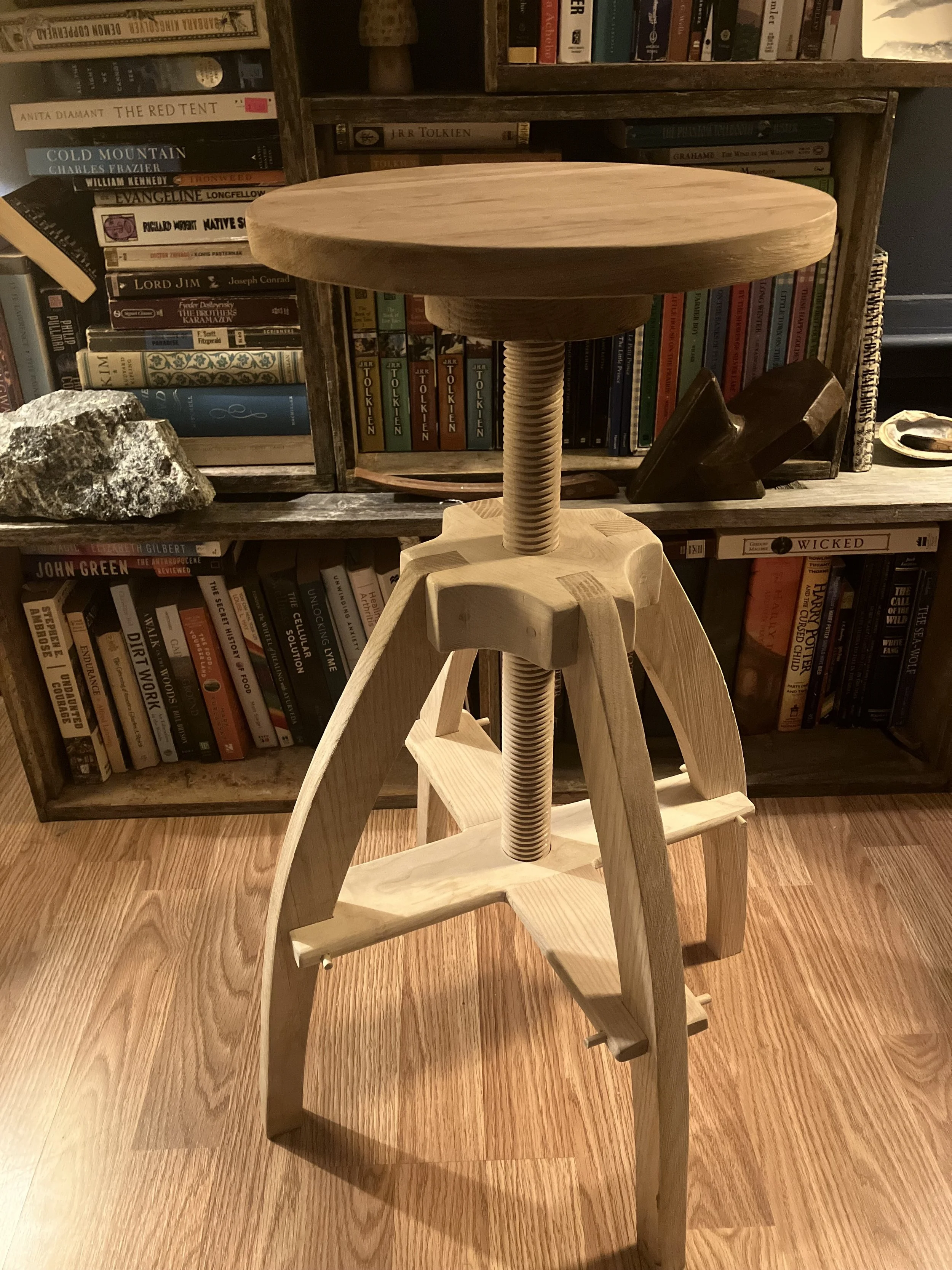 A wooden stool with a round top, supported by a threaded metal screw in the center, standing on a wooden floor in front of a bookshelf filled with books.