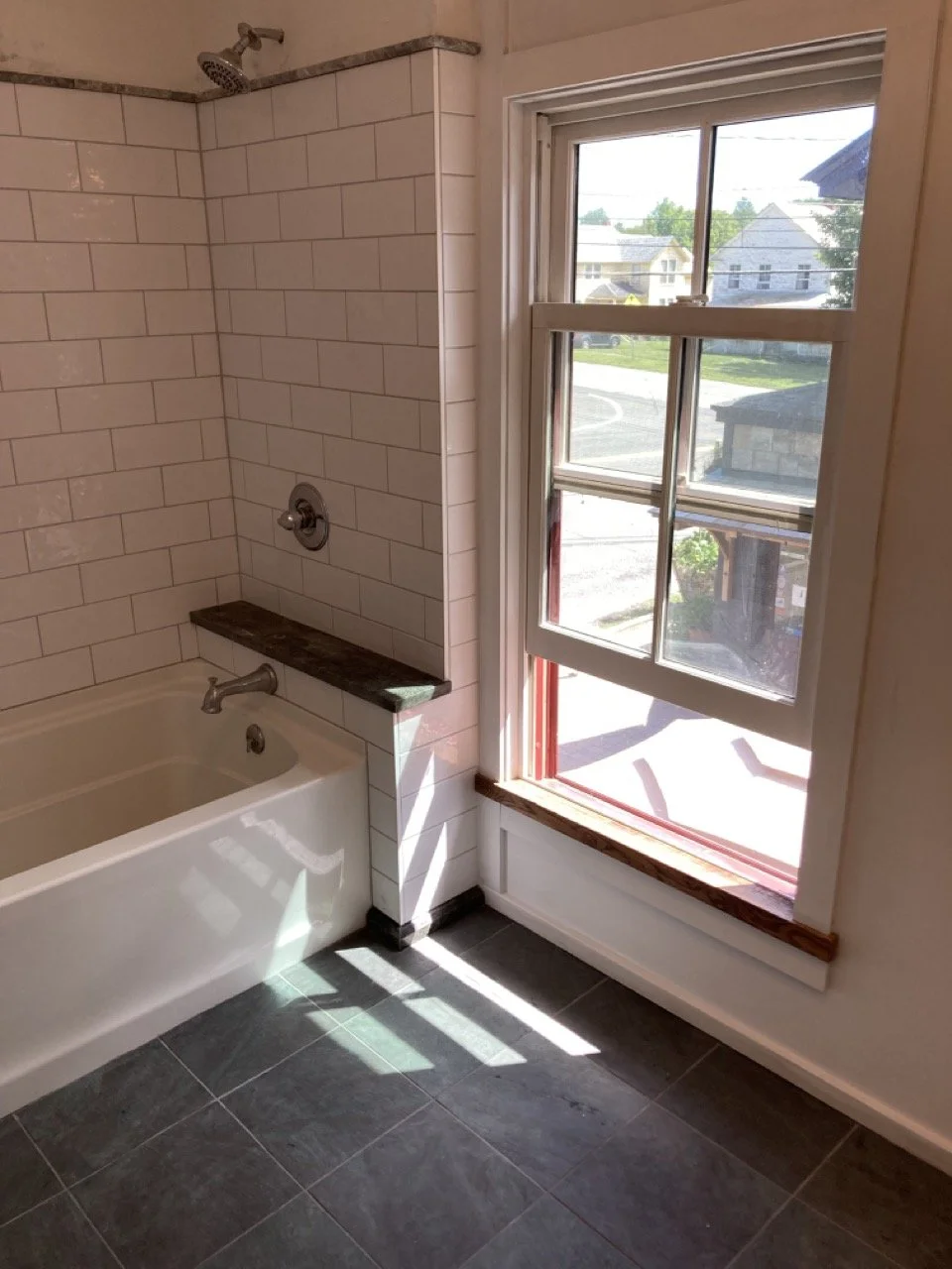 Bathroom with white subway tile walls, a bathtub, a large window with a wooden sill, and a view of neighboring houses outside.