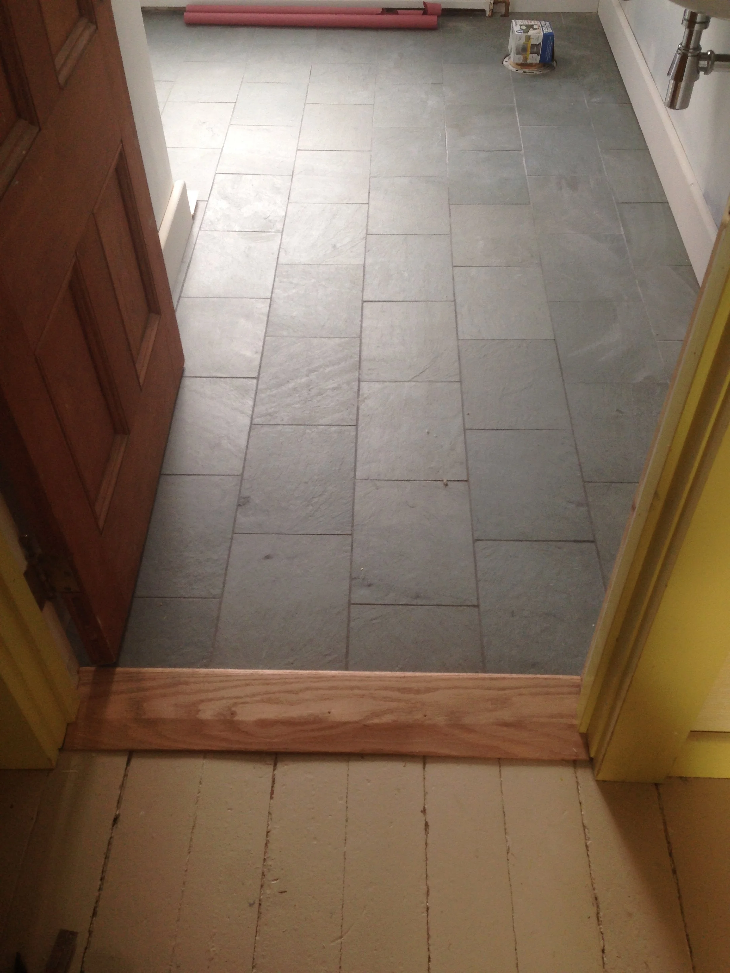 Interior view of a doorway leading into a tiled room with gray slate tiles, a wooden door on the left, a yellow wall on the right, and construction materials at the back.