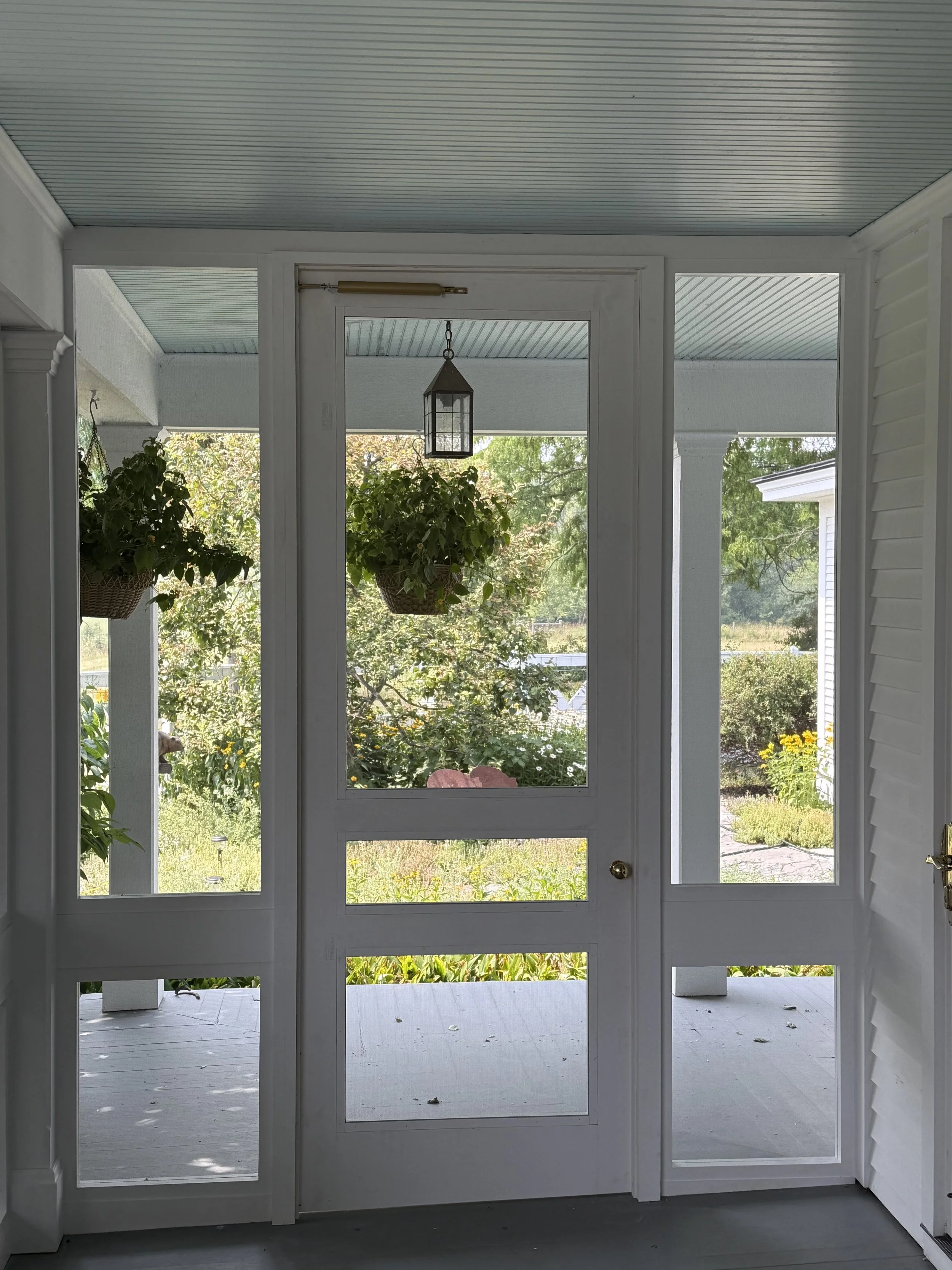 Front door with glass panels leading to a porch with hanging plants and a view of a garden with green trees and yellow flowers.