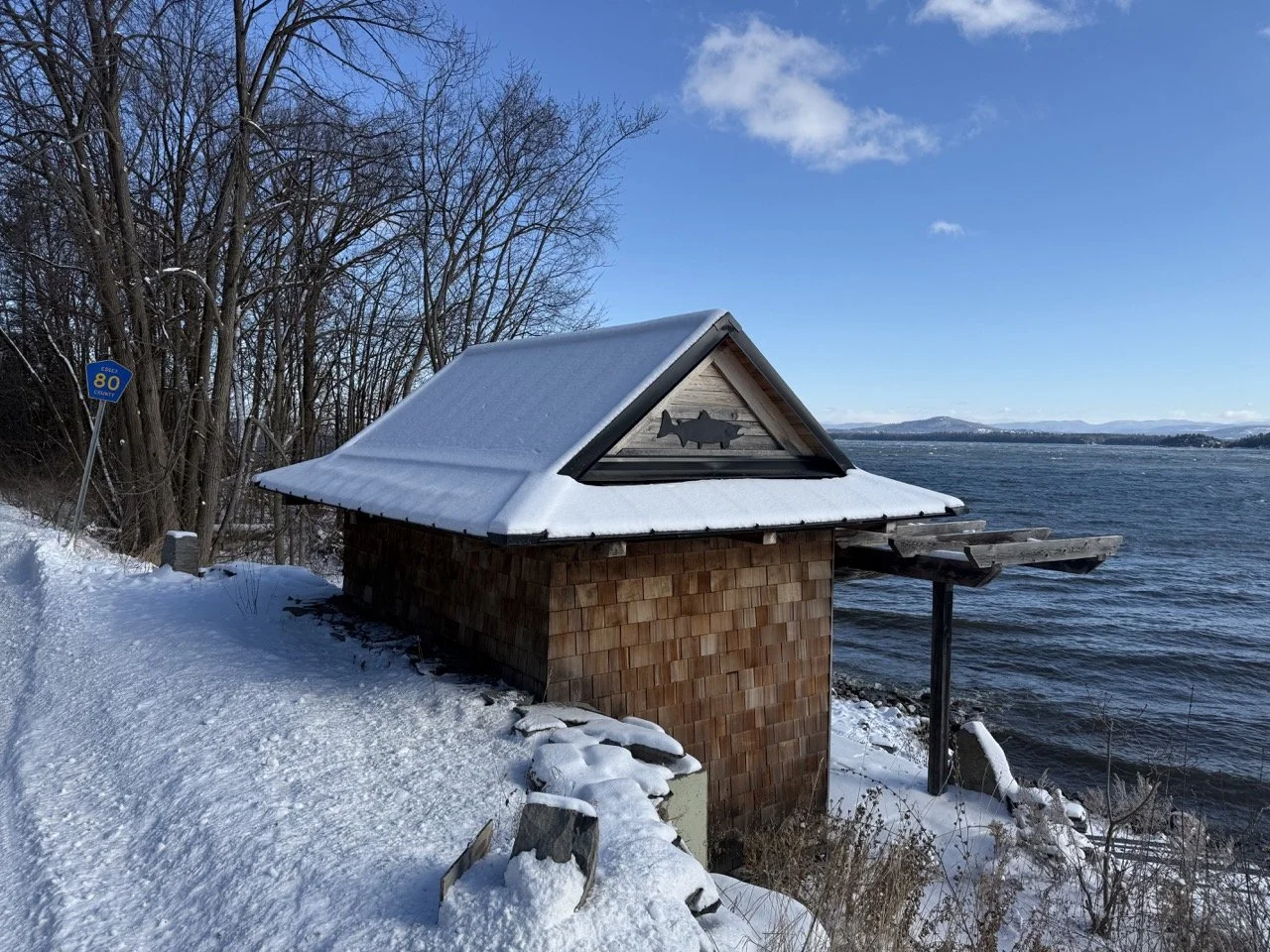 A small wooden boathouse with a snow-covered roof situated along a lake, with leafless trees and a clear blue sky in the background.