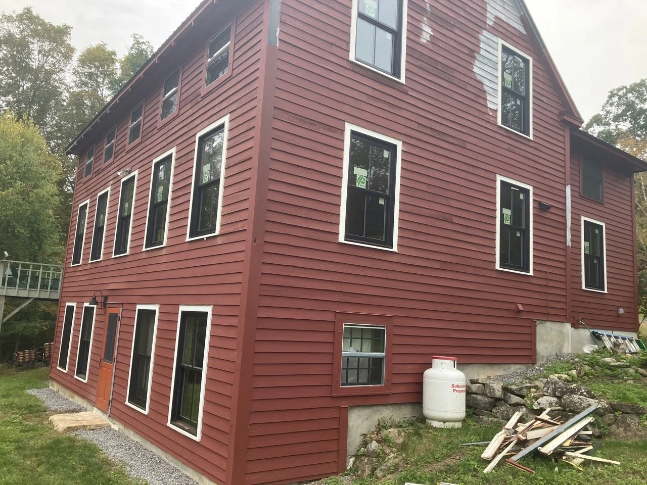Three-story red house under construction with multiple black-framed windows, a propane tank, and construction debris on the grass.