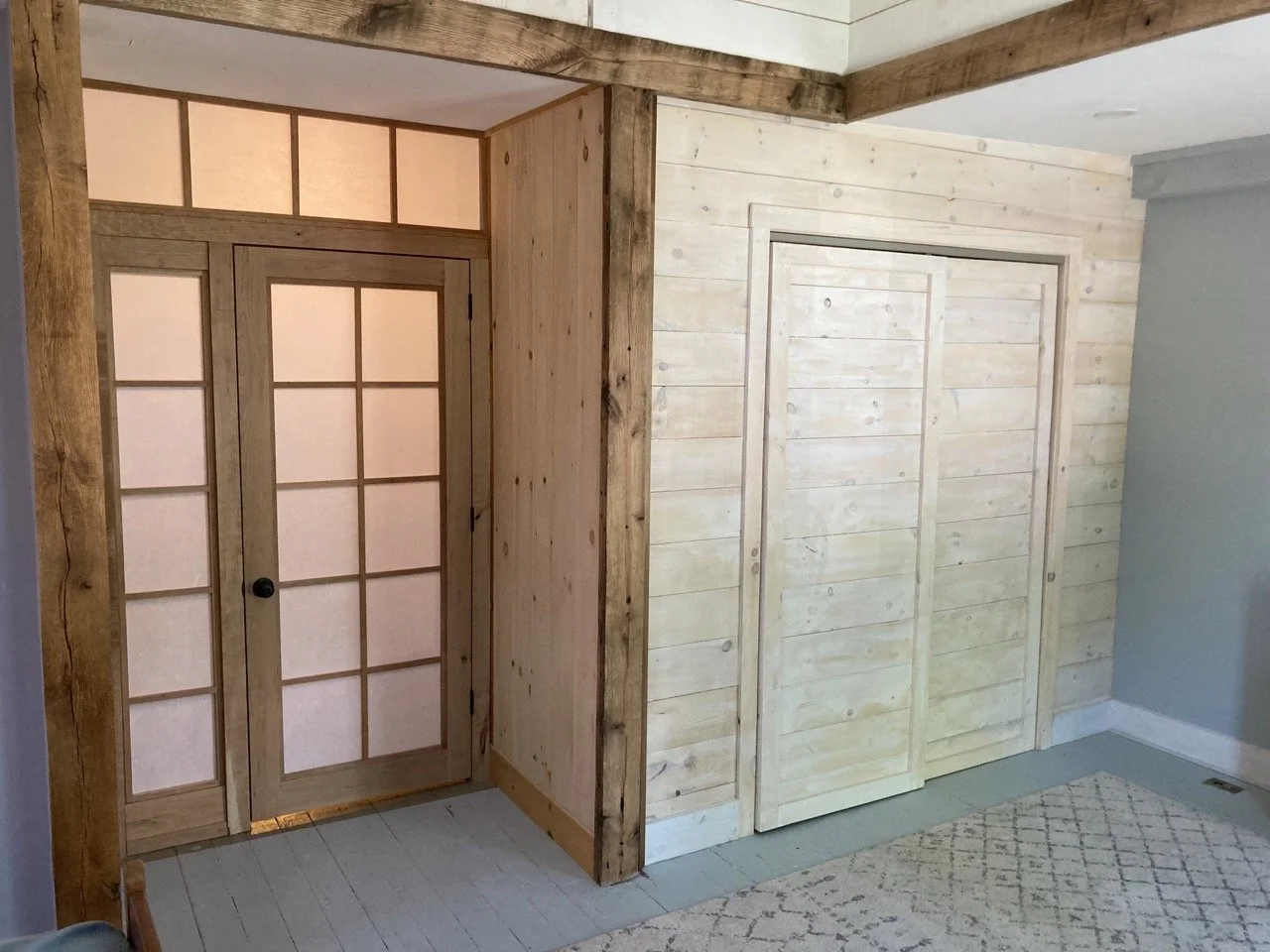 Interior of a room with wooden walls and doors, including a glass-paneled door and a sliding closet door, with a section of carpeted floor and a small area of painted floor.