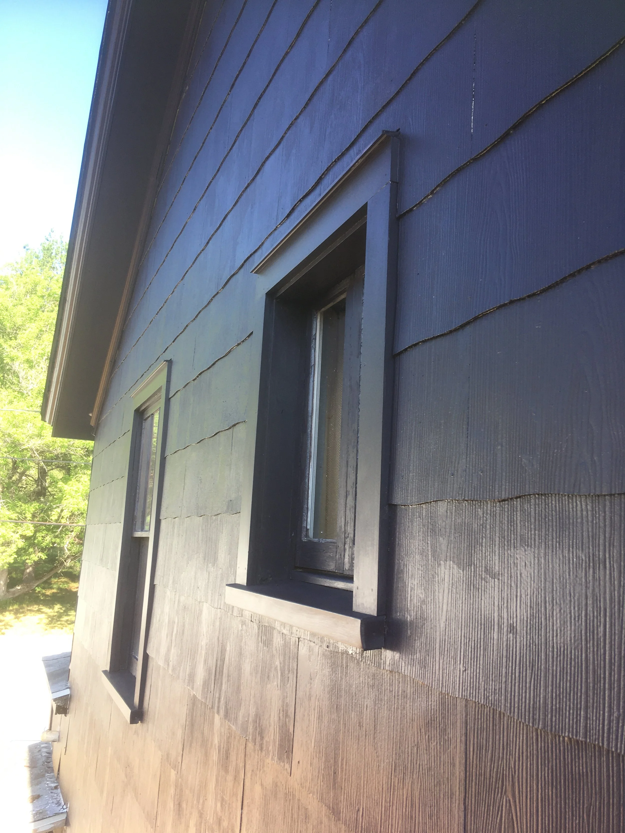 Close-up of a house's exterior wall with dark horizontal siding, showing two vertically oriented rectangular windows with black trim and window sills, under a sloped roof.