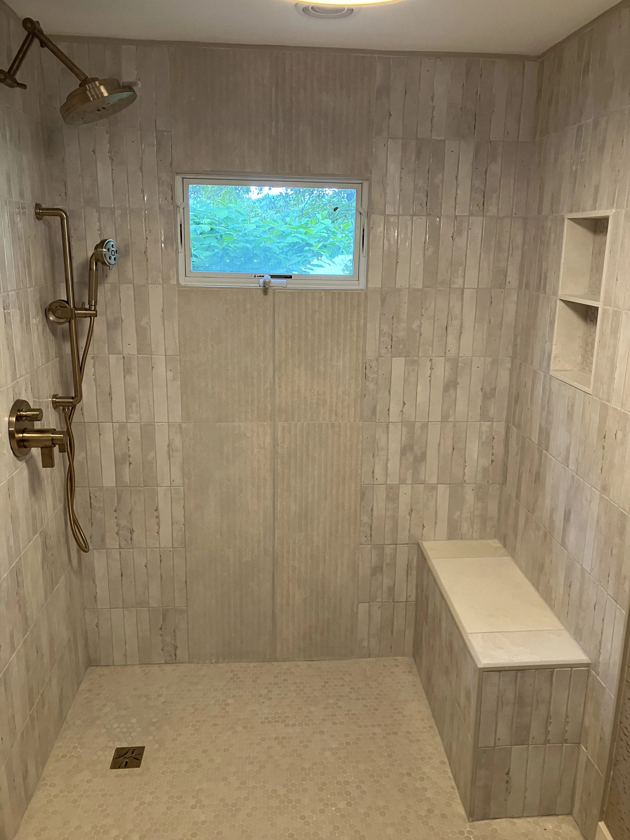 A walk-in shower with beige tiled walls and floor, a window showing green foliage outside, a built-in bench, and a showerhead with a handheld attachment.