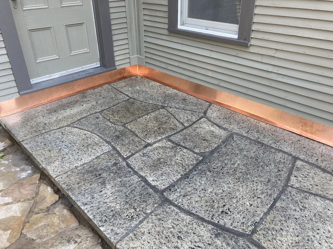 Front porch with a stone paver walkway, new copper metal flashing at the base of the house near the door and window, beige siding and a gray door.