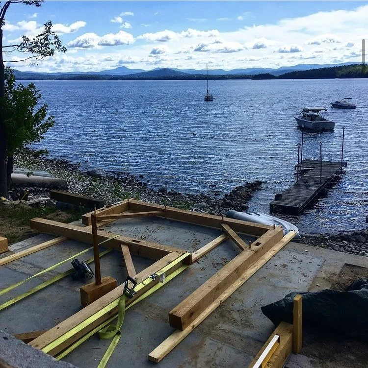 A lakeside scene with a partly constructed wooden dock and tools in the foreground, calm water with boats, and distant mountains under a partly cloudy sky.