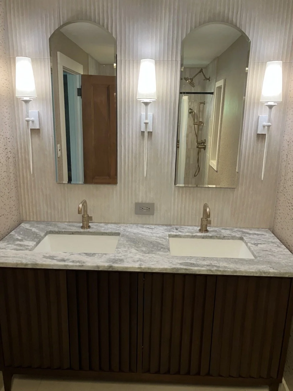 Bathroom vanity with two sinks, marble countertop, and two mirrors. Wall-mounted sconces with white shades are on either side of the mirrors. A walk-in shower with brass fixtures is reflected in the right mirror.