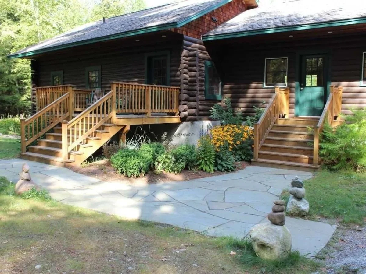 A log cabin with two front porches, each accessed by wooden stairs, surrounded by greenery and colorful flowers, with a stone pathway leading up to the stairs.