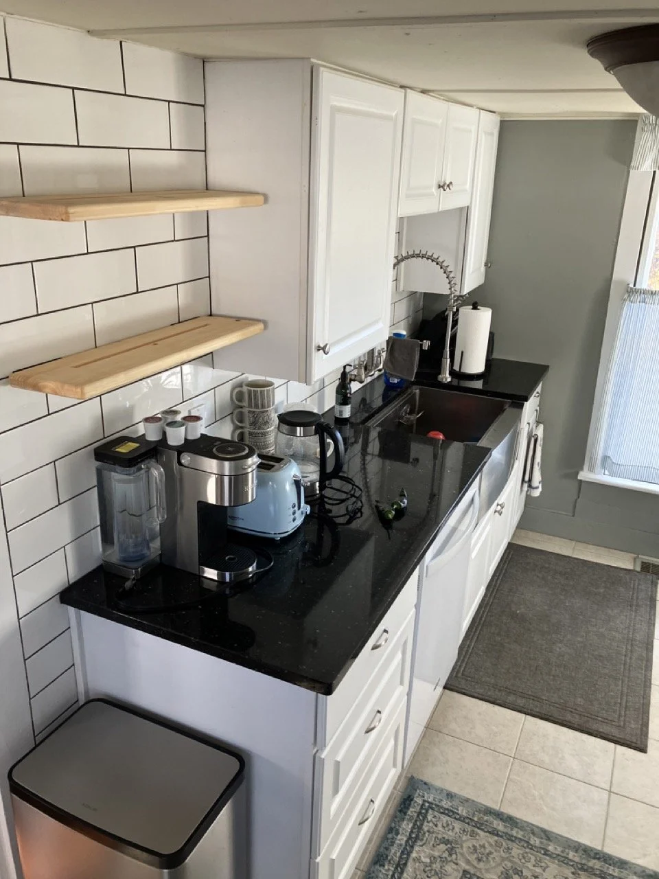 A compact kitchen with white cabinets, black granite countertop, and white tile backsplash with black grout. There are wooden floating shelves on the wall. On the counter are various small appliances including a coffee maker, blender, and toaster, al