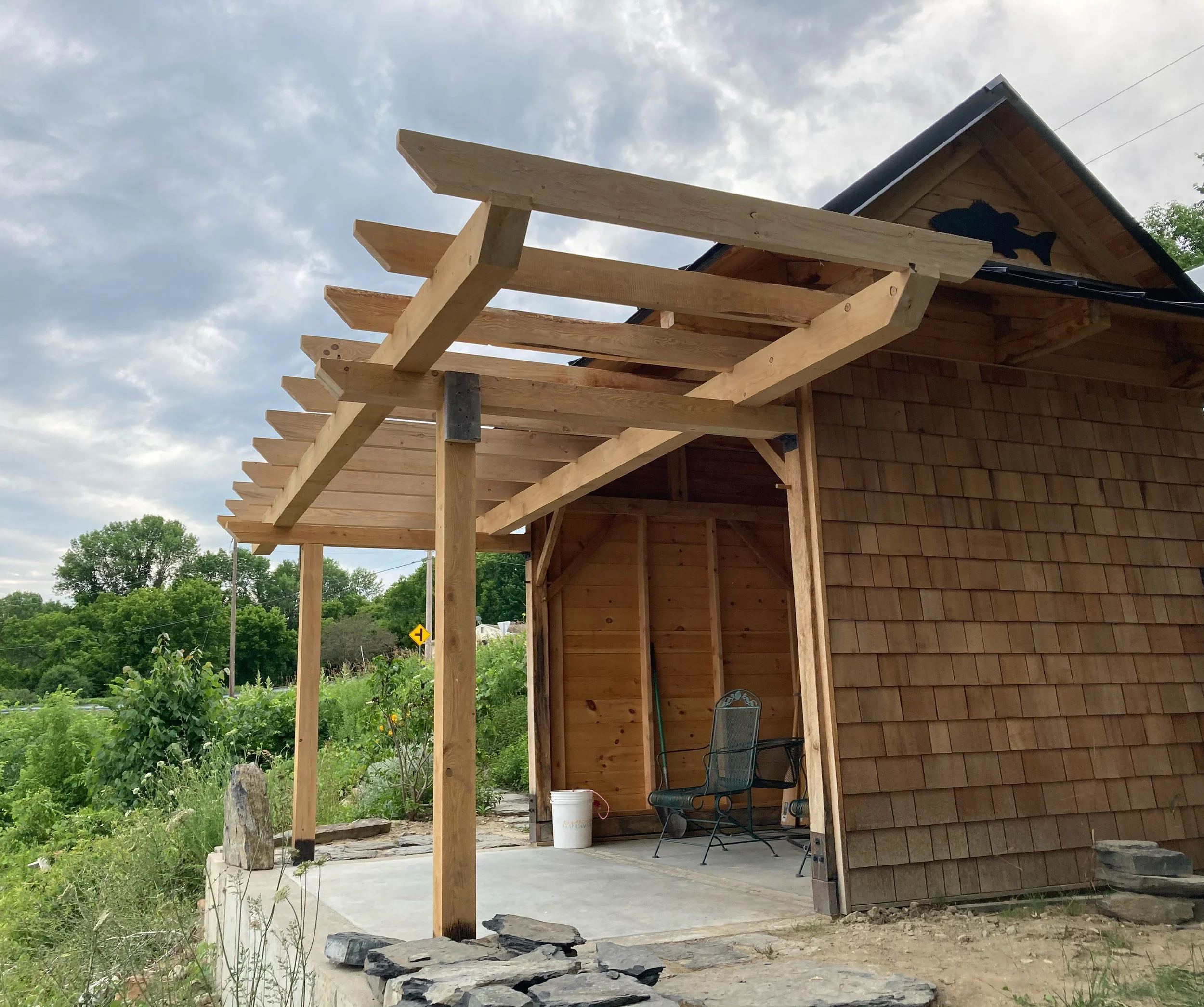 Construction of a new porch or overhang attached to a wooden house, with timber beams, a concrete base, and outdoor furniture, on a cloudy day.
