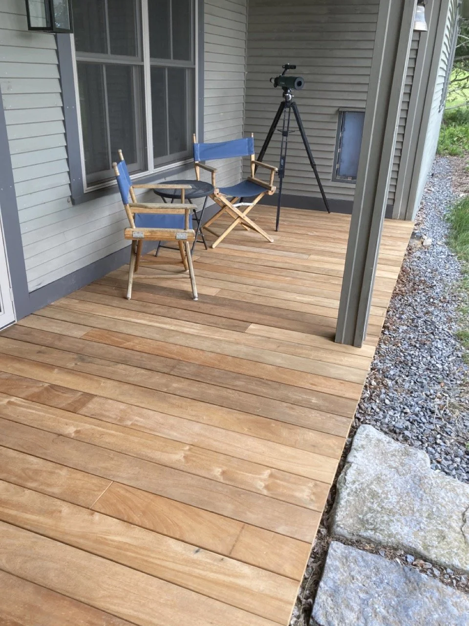 A porch with wood decking. There are two blue and wood folding chairs, a small black table, and a telescope on a tripod. The porch has siding and a window, with grass and gravel outside.