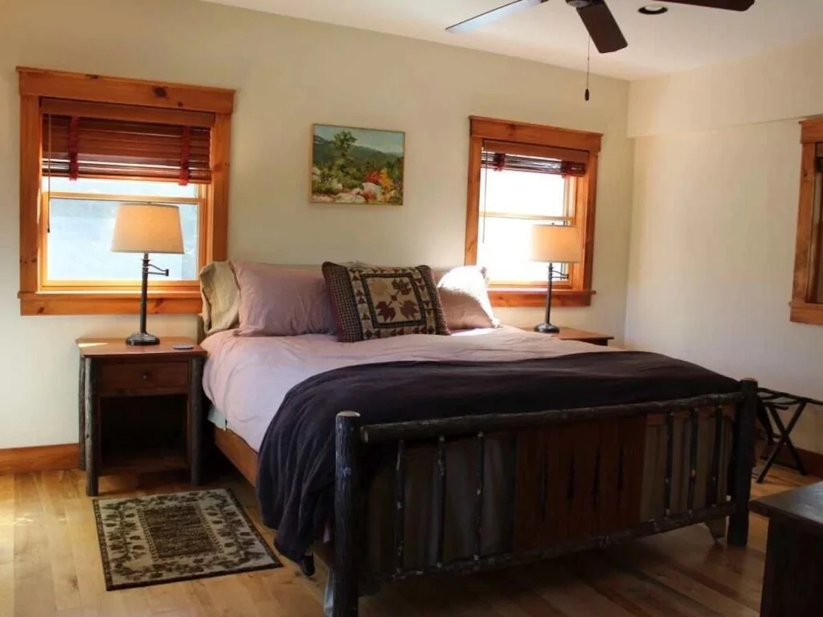 A bedroom with a rustic style, featuring a wooden bed frame with beige and brown bedding, two bedside tables with lamps, two windows with wooden blinds, a small painting on the wall, and hardwood flooring.