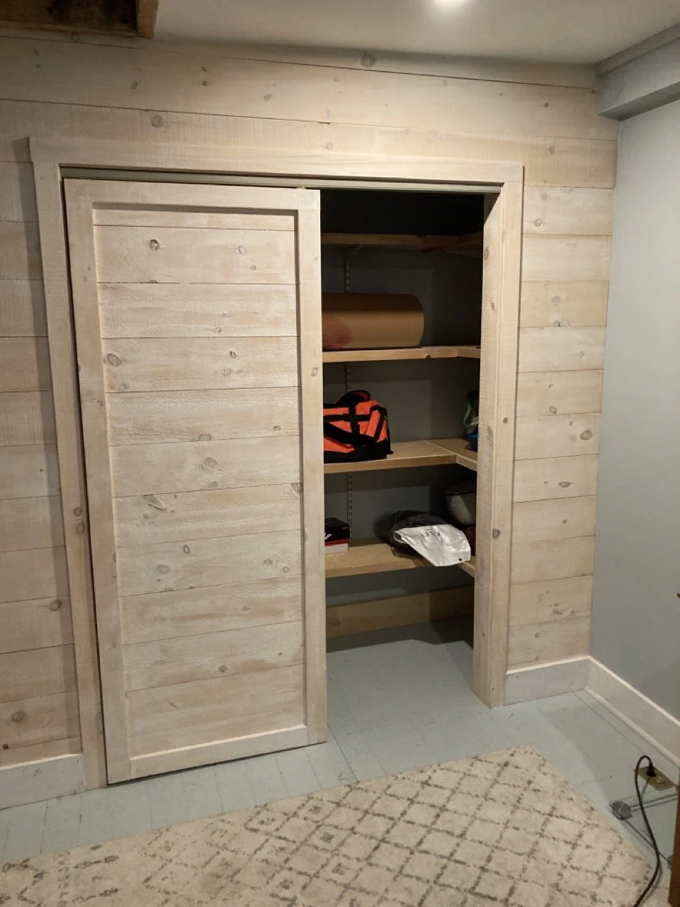 Open wooden closet with shelves containing a paper roll, orange and black bag, wrapped item, and other miscellaneous items.