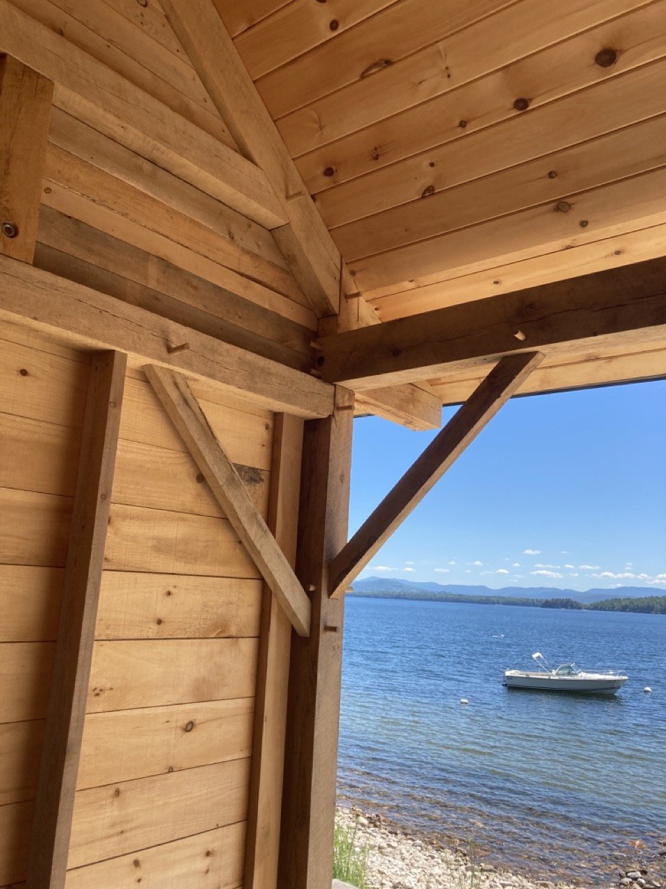View of a lake with a boat, seen from inside a wooden structure, exposed wood framed walls and roof, on a sunny day with a clear blue sky.