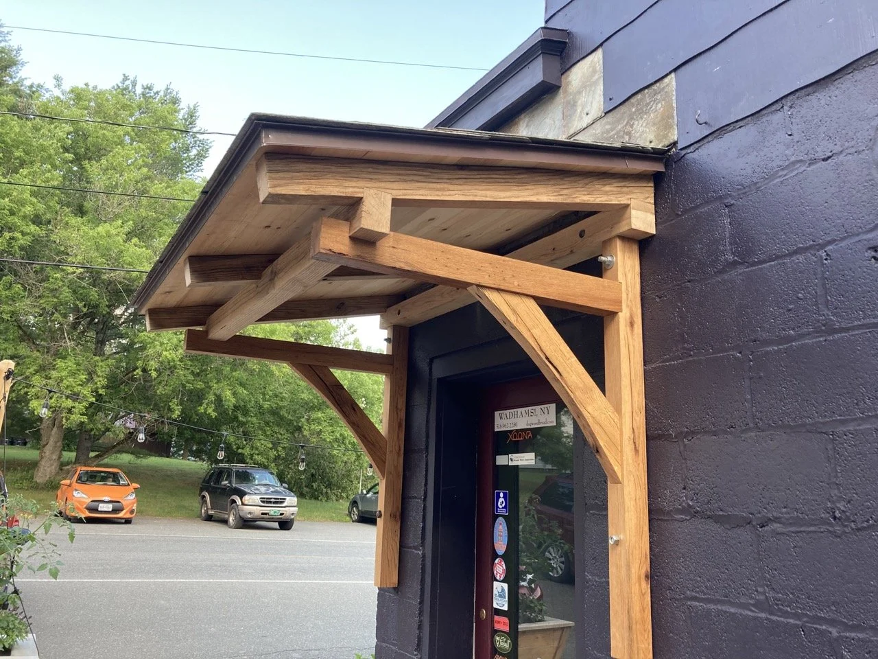 Close-up of a small wooden awning above an entrance door of a building with black painted brick wall, with parked cars and trees in the background.