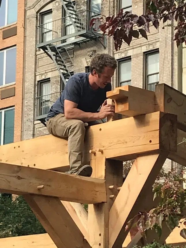 A man sitting on a large timber framed structure,  fitting joinery together outdoors in front of a building with fire escapes.