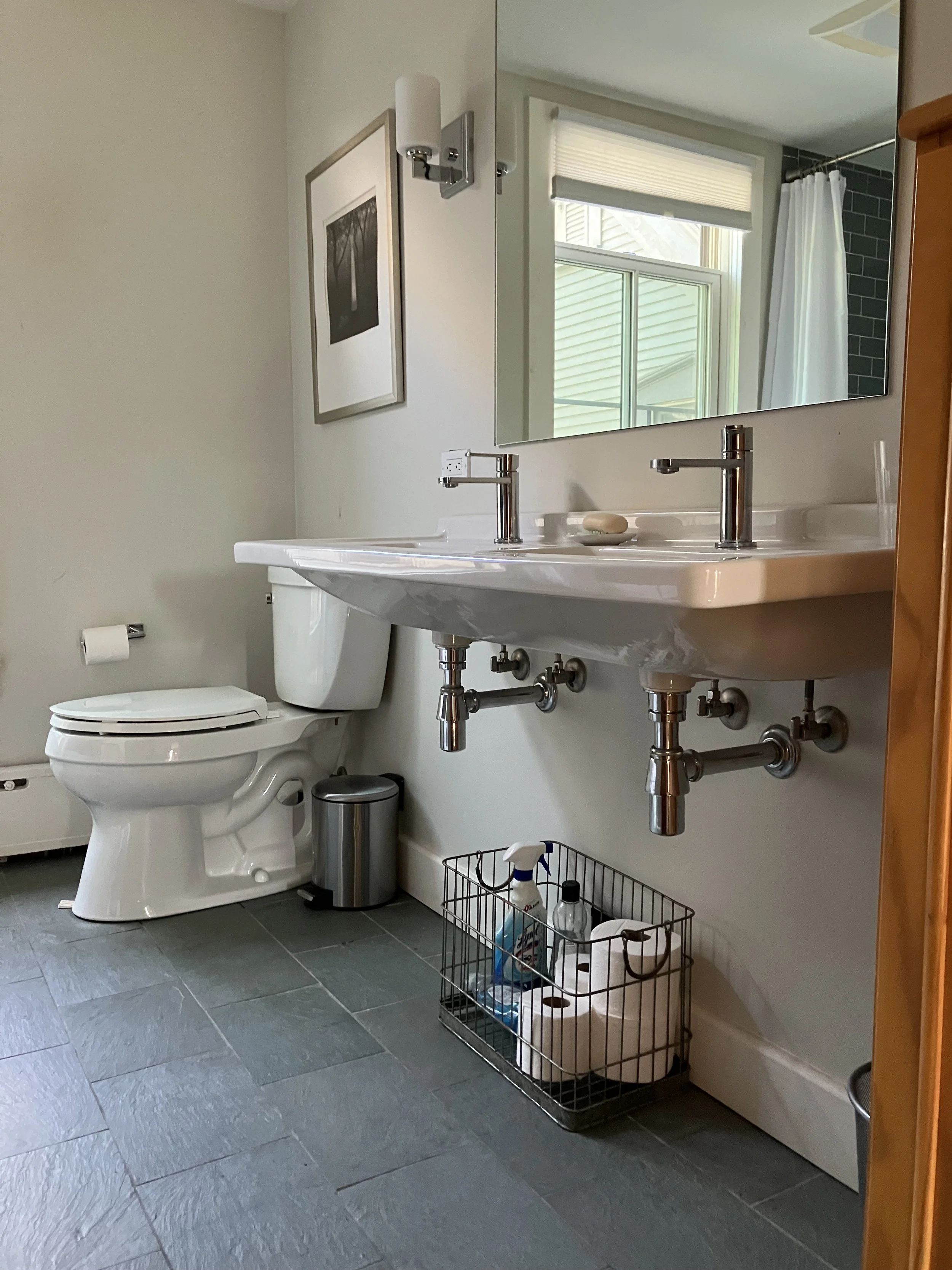 A bathroom with a white toilet, a wall-mounted mirror, and a white sink with two faucets. There is a small framed picture on the wall, a window with a blind, and a green tiled shower curtain in the background. The floor is tiled with large gray tiles, and there is a wire basket with cleaning supplies under the sink.