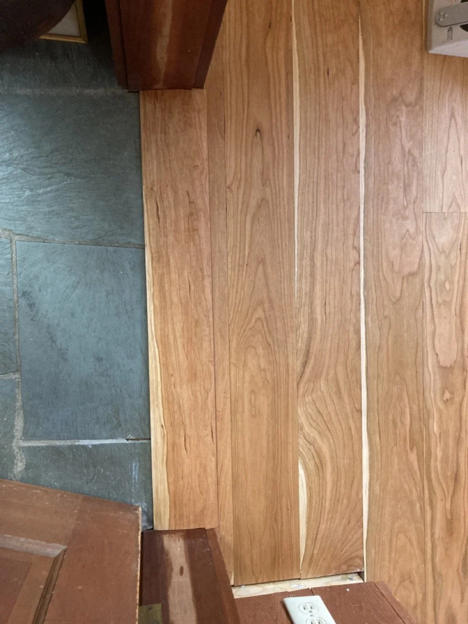 Wooden floor transition strip between stone and wood flooring near electrical outlet.