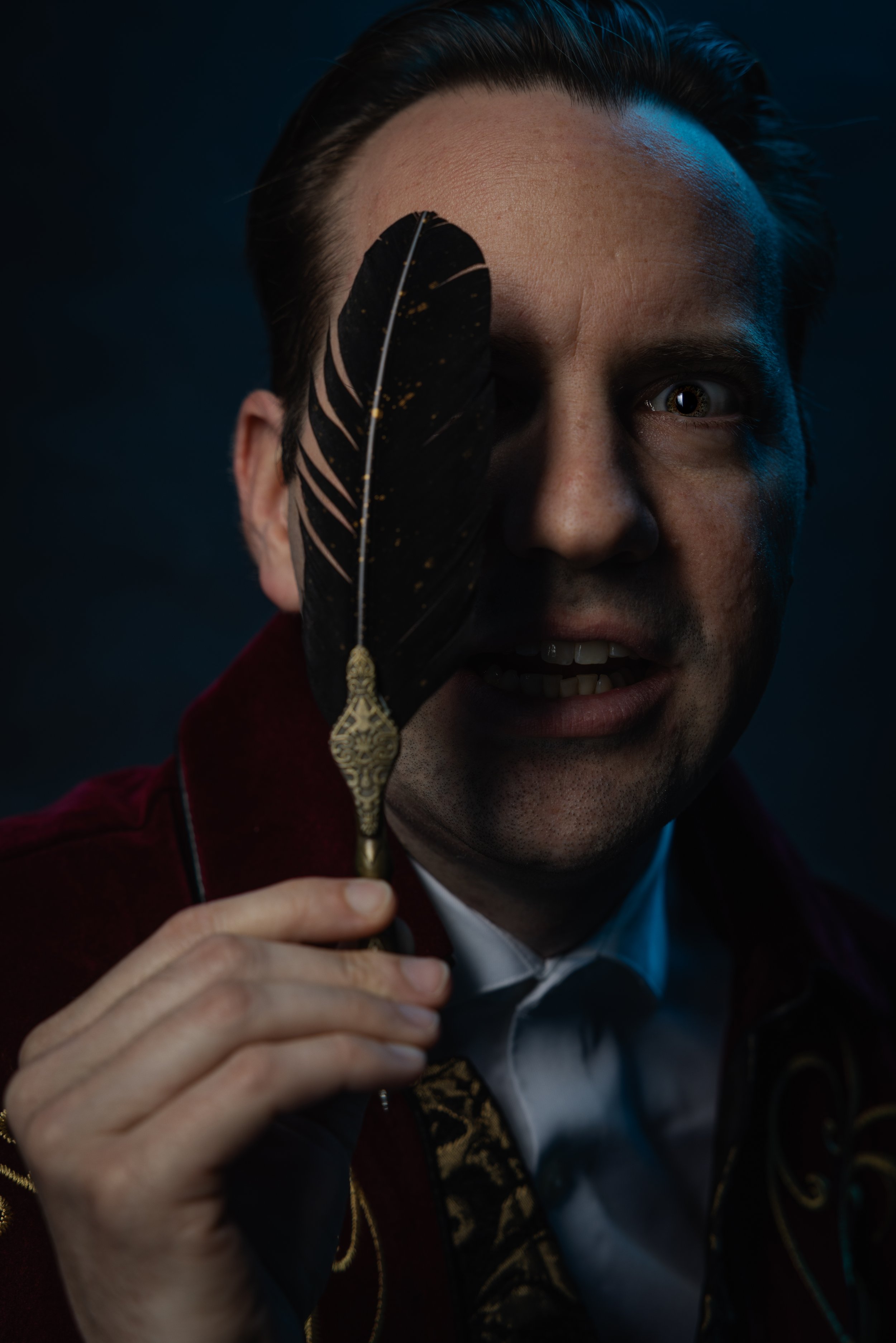 A man holds a black feather with gold accents in front of his face, obscuring one eye, in a dark setting.