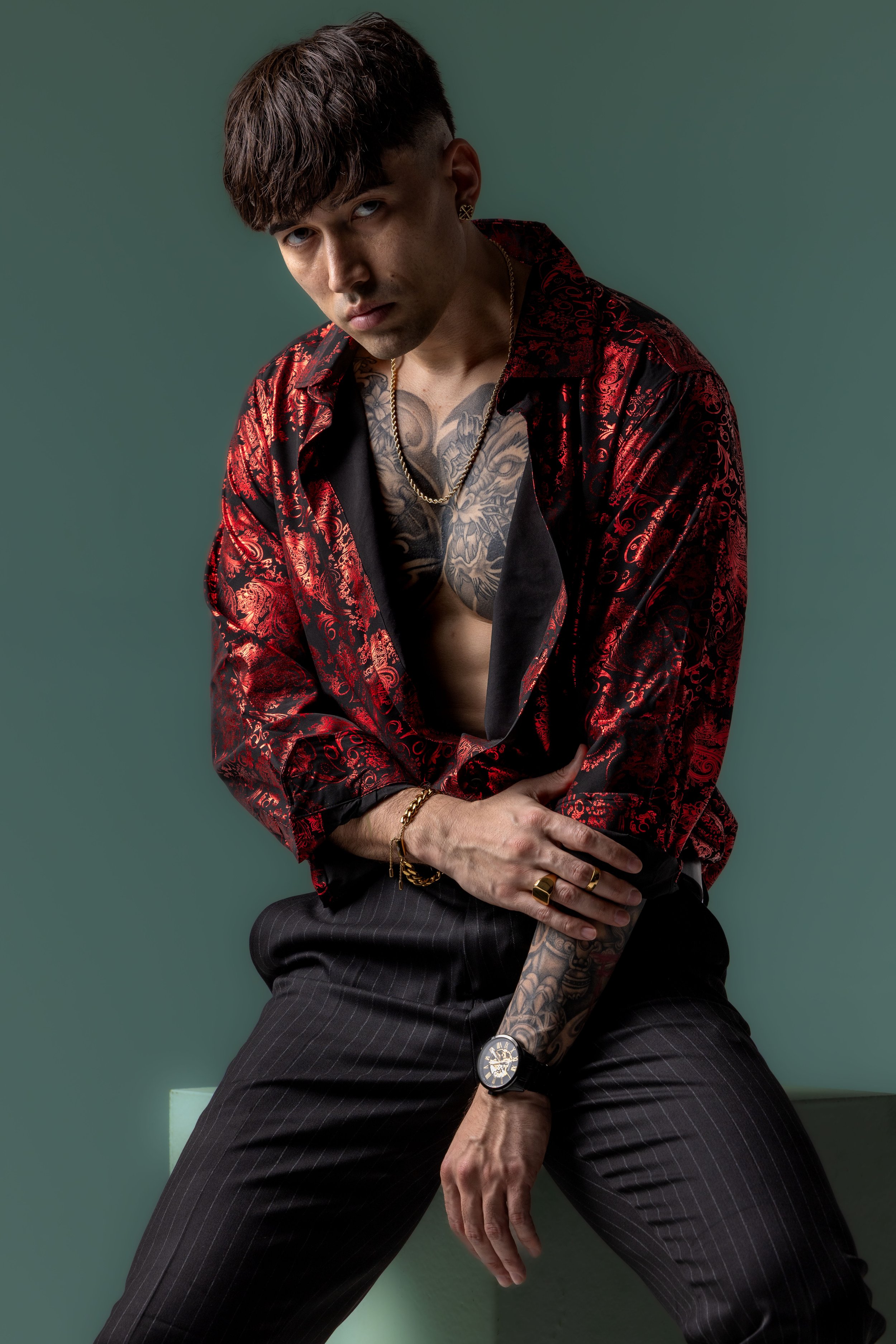 A young man with tattoos and dark hair, wearing a black and red patterned silk shirt partially open, black pinstripe pants, gold jewelry, and a wristwatch, sitting against a plain green backdrop.