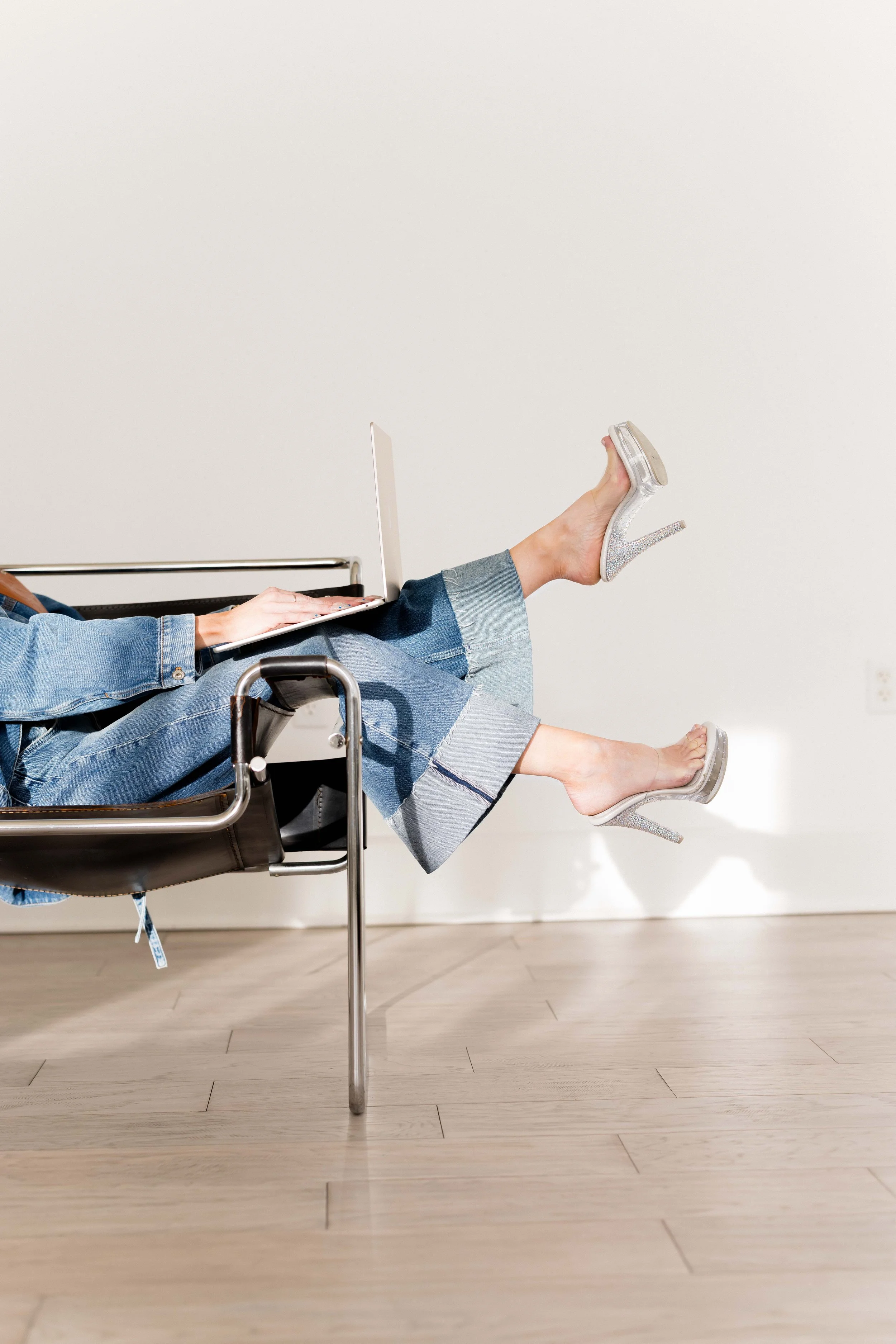 Person sitting in a chair with legs extended, wearing jeans and glittery high heels, working on a laptop.