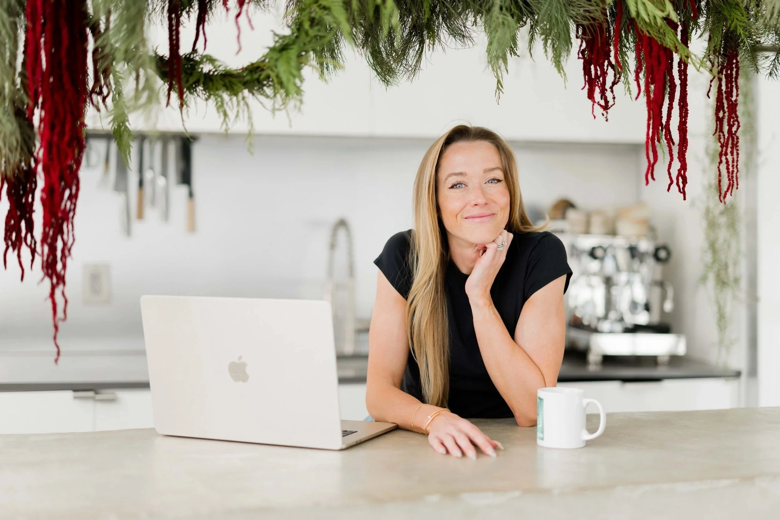 A woman sitting at a kitchen counter with a MacBook laptop and mug, smiling, with decorative hanging greenery and red accents overhead.