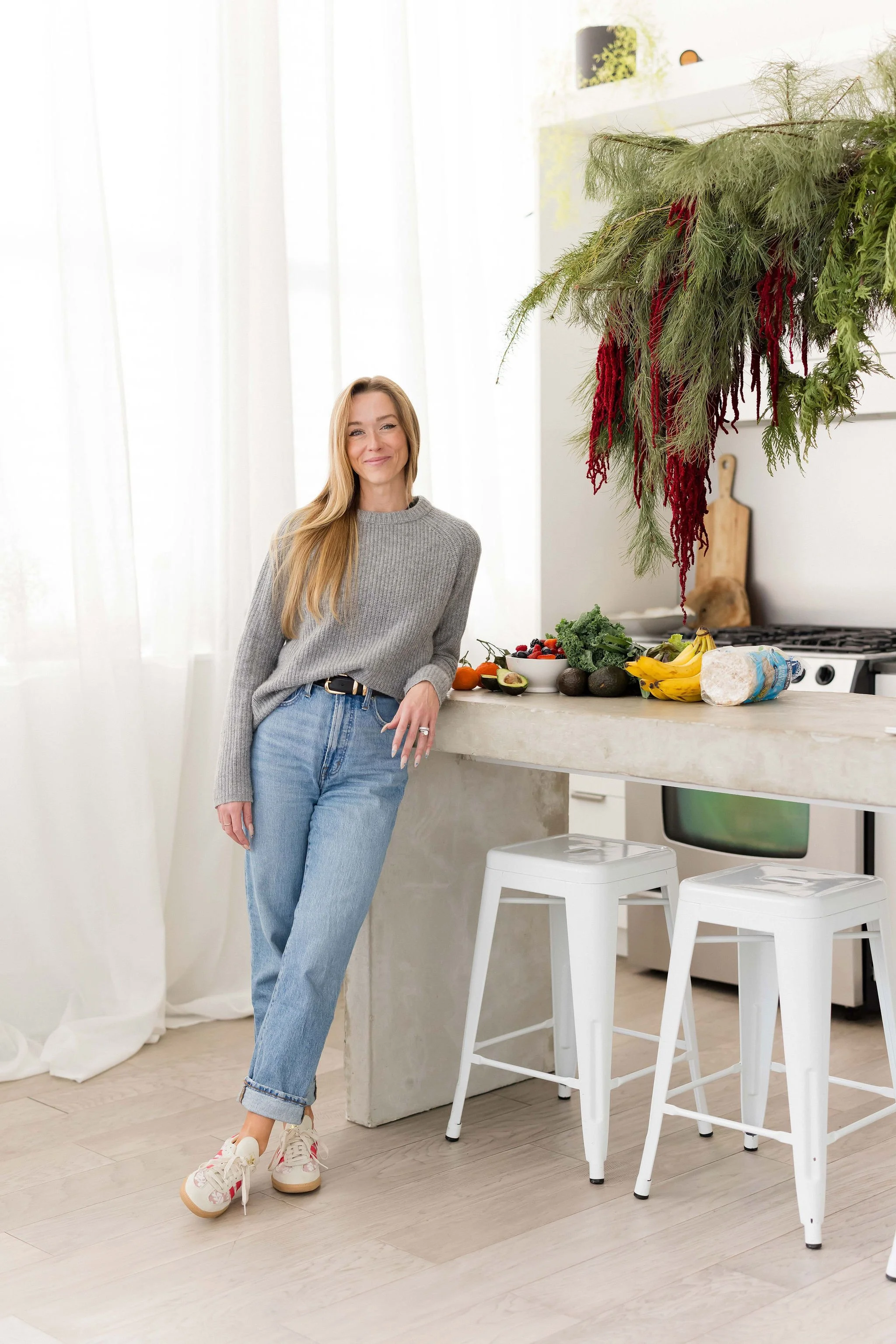 A woman with long blonde hair leaning against a kitchen counter, smiling, in a modern kitchen with white curtains and natural light. There are fruits and vegetables on the counter and hanging greenery overhead.