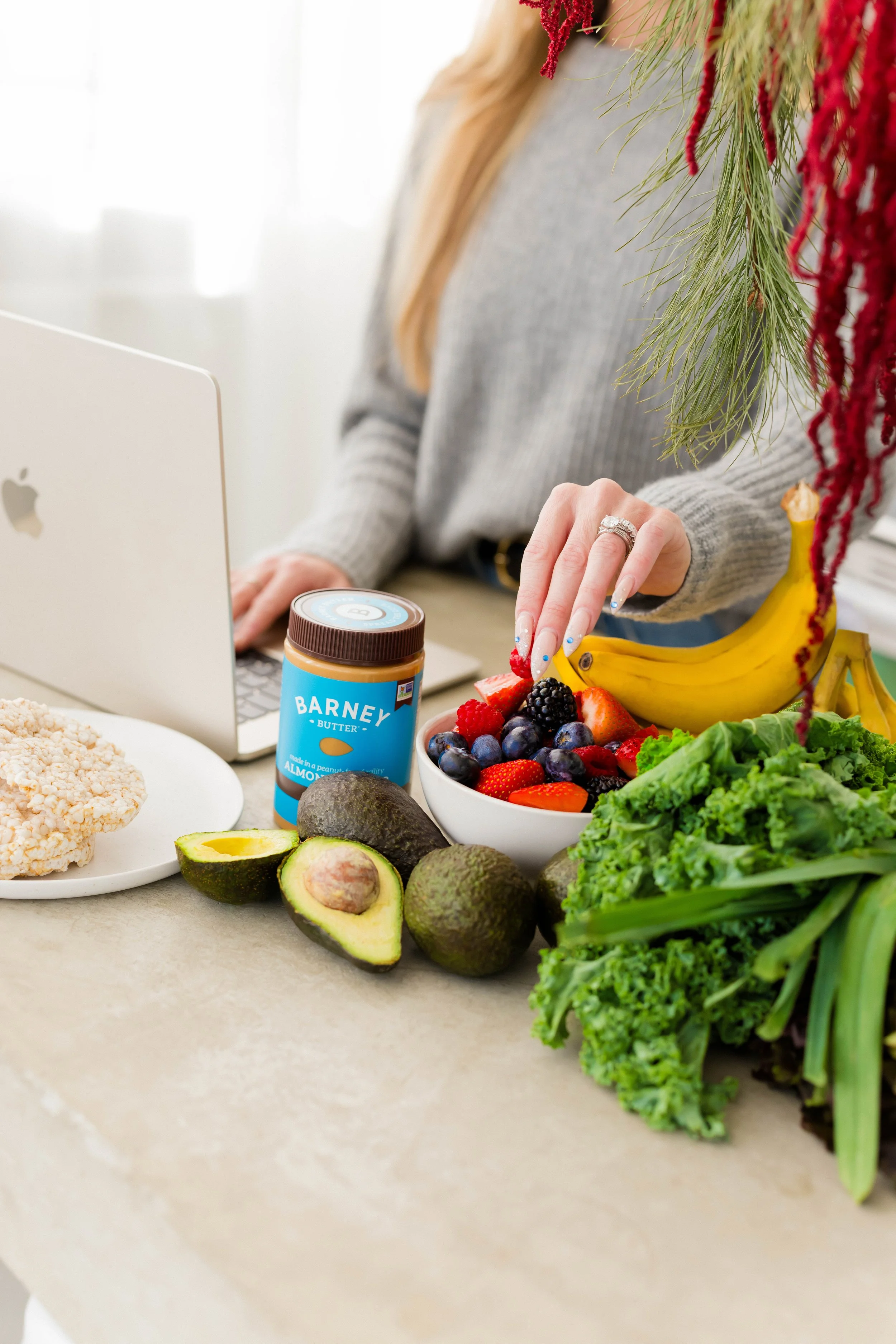 A woman preparing a healthy fruit snack with strawberries, blackberries, blueberries, bananas, avocados, gardening greens, rice cake, and almond butter on a kitchen counter.