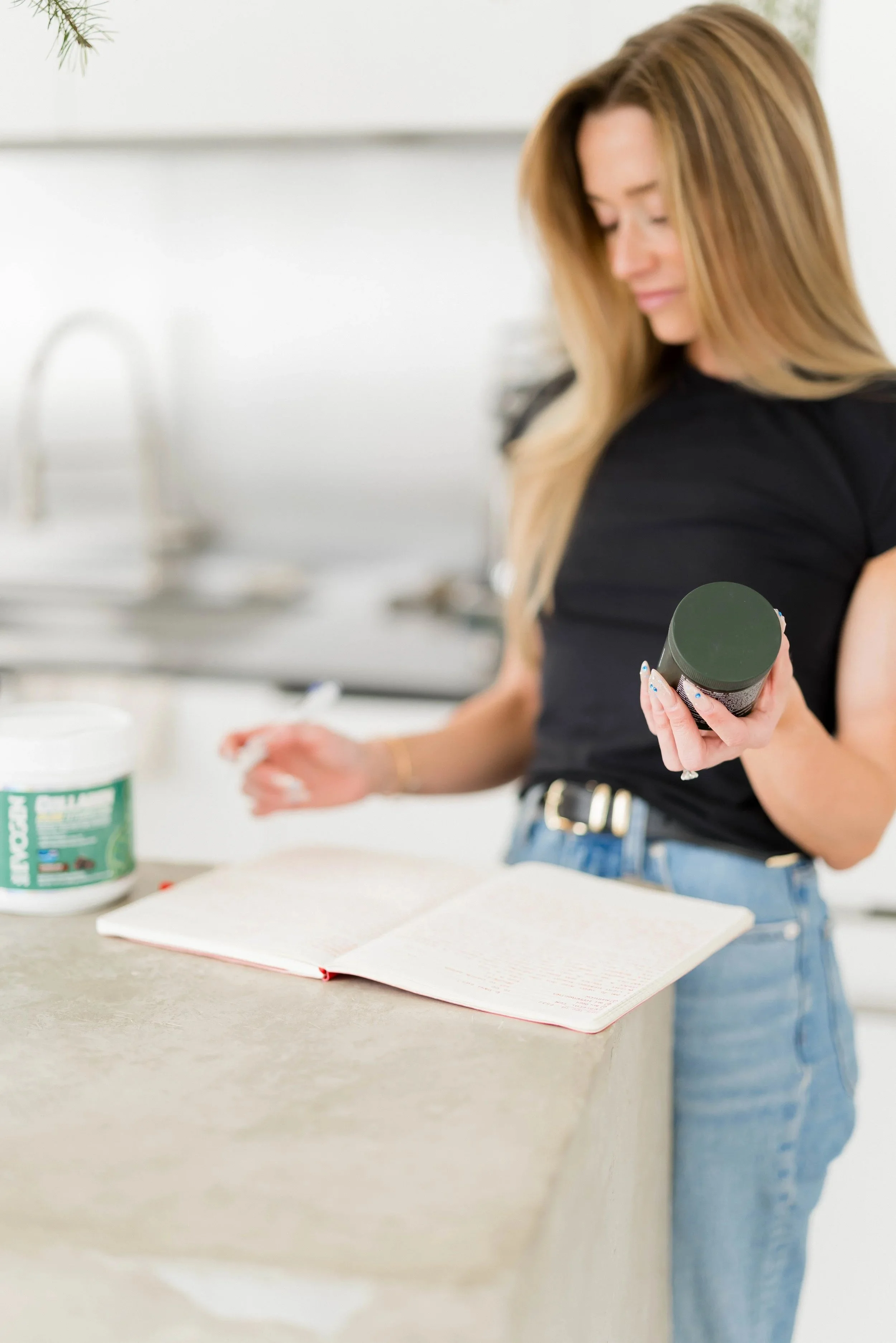 A woman standing in a kitchen, holding a container of supplements and an open book on the counter.