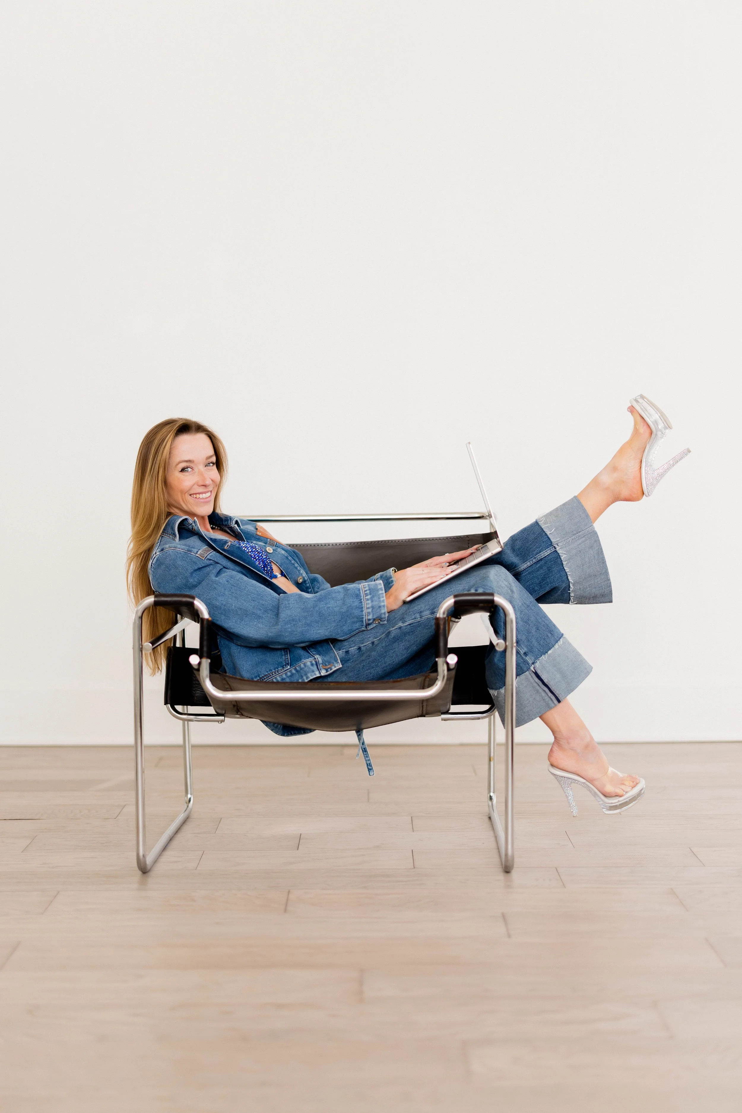 A woman in a denim jacket and jeans with sparkly high heels sitting on a modern black chair, smiling while using a laptop with her legs raised and crossed, against a plain white wall and wood floor background.