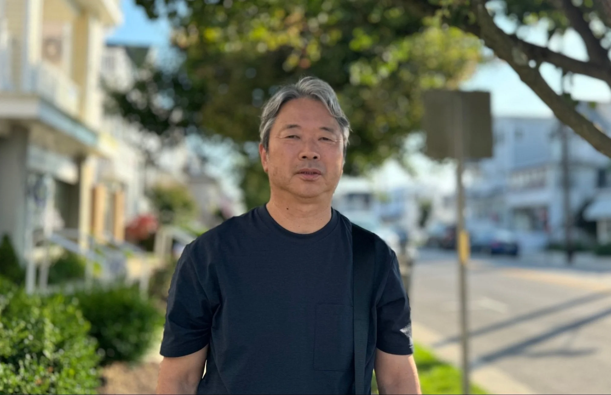 A middle-aged man with gray hair standing outdoors on a sunny day, with trees and residential houses in the background.