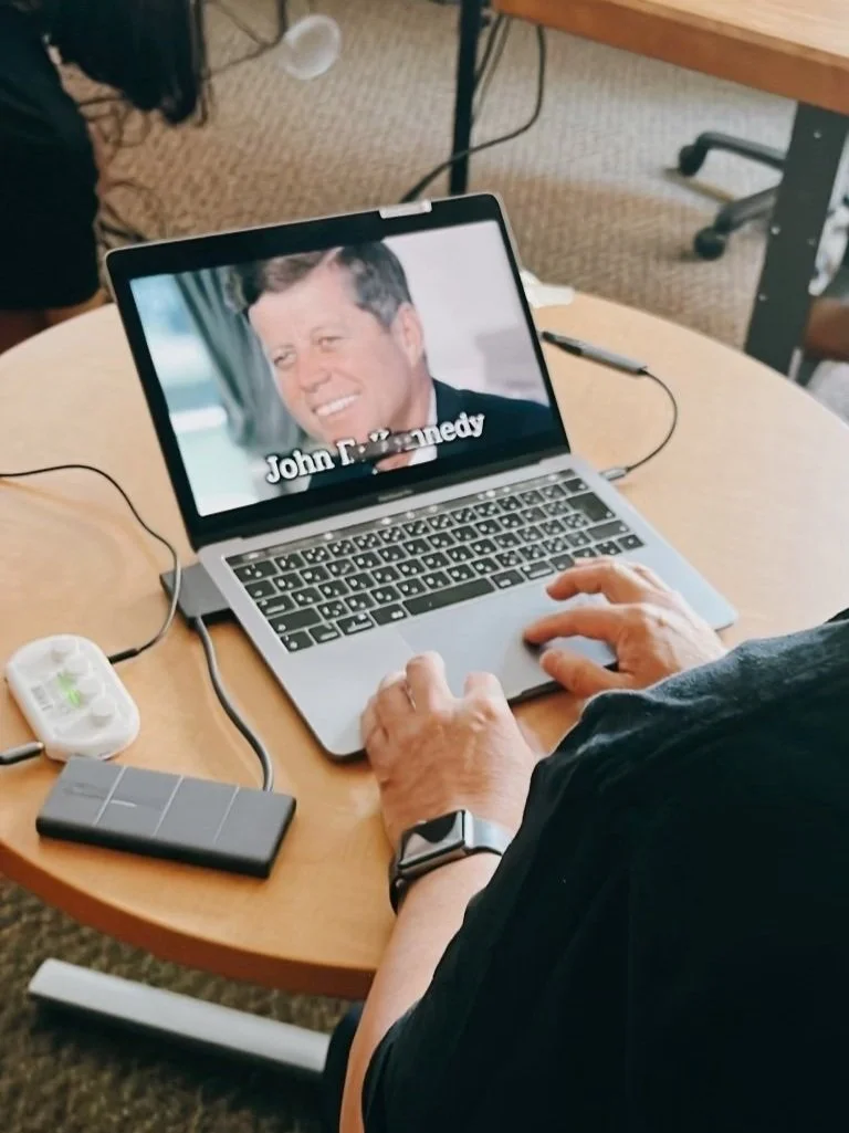 A person sitting at a round wooden table watching a video of a man with the name 'John D. Kennedy' on a laptop screen.