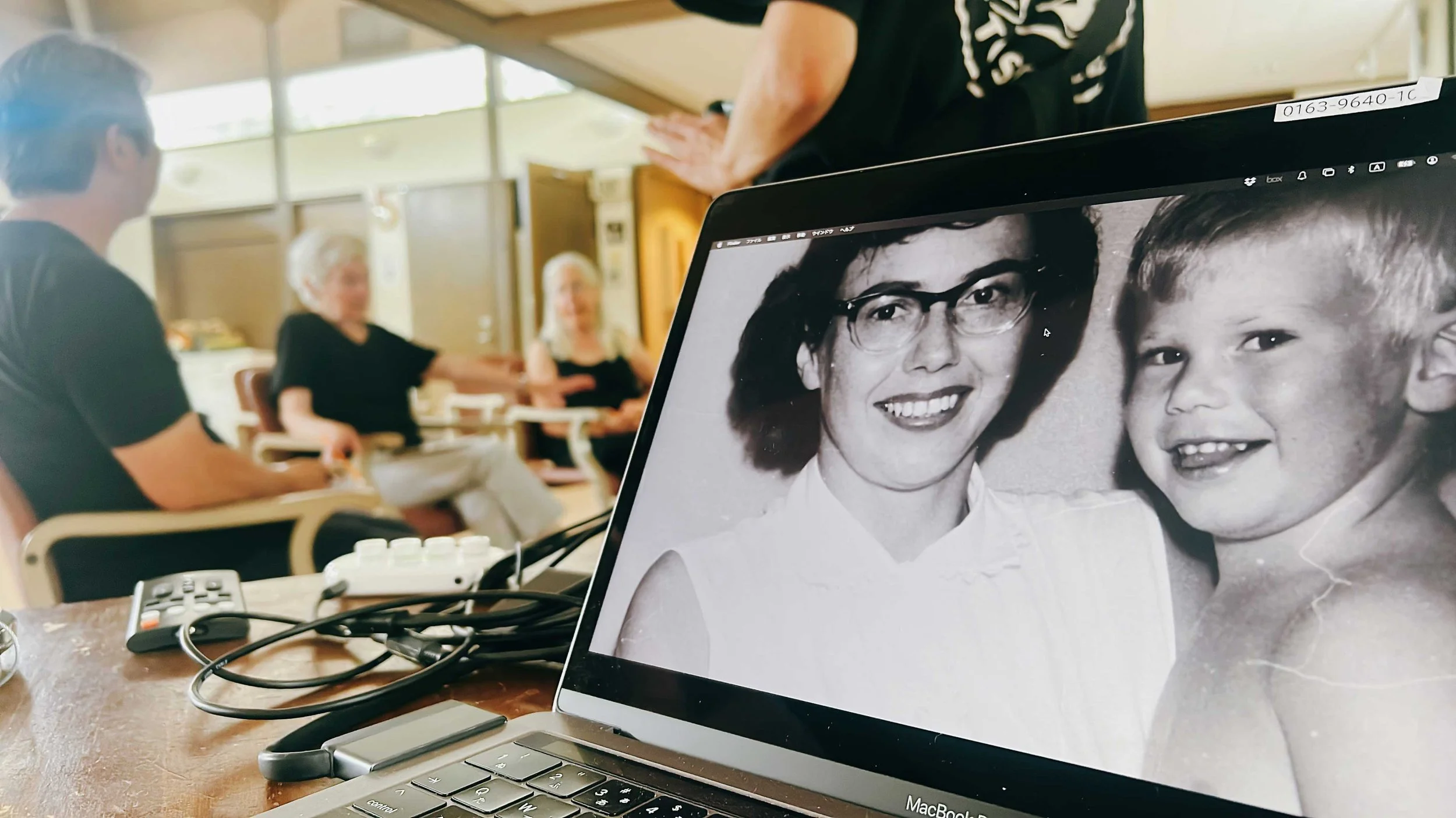 A laptop displaying a black and white photo of a woman with glasses and a young boy smiling. In the background, three people are sitting and talking in a room with large windows and wooden doors.