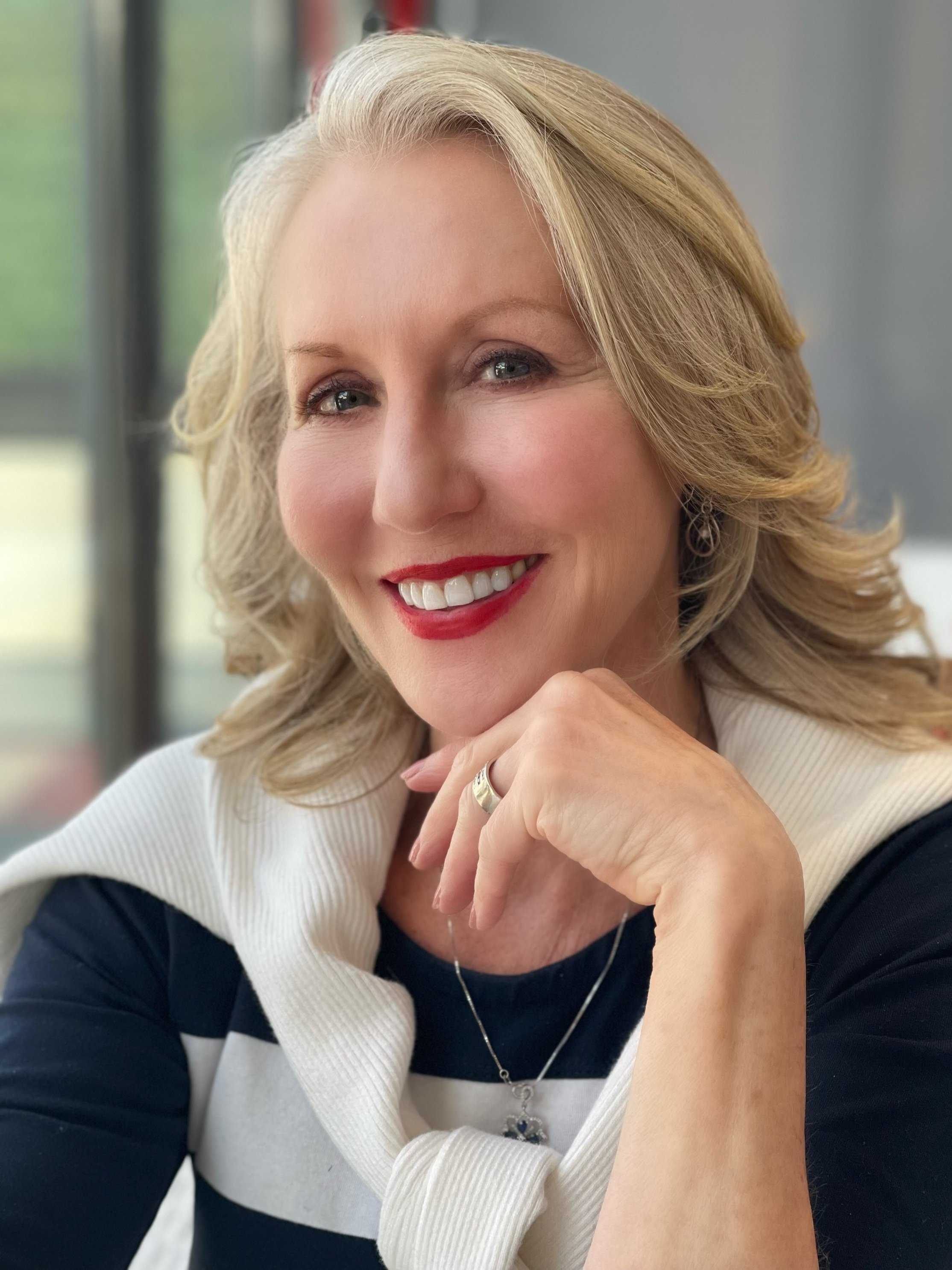 Close-up portrait of a smiling middle-aged woman with blonde hair, red lipstick, and blue eyes, wearing a navy blue and white top with a cream sweater draped over her shoulders, sitting indoors near a window.