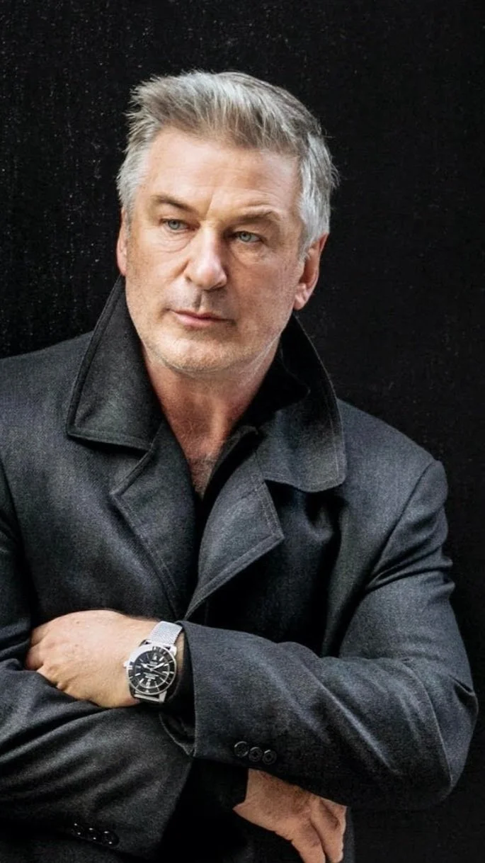 A middle-aged man with gray hair and blue eyes posing with arms crossed, wearing a dark gray coat and a wristwatch against a black background.