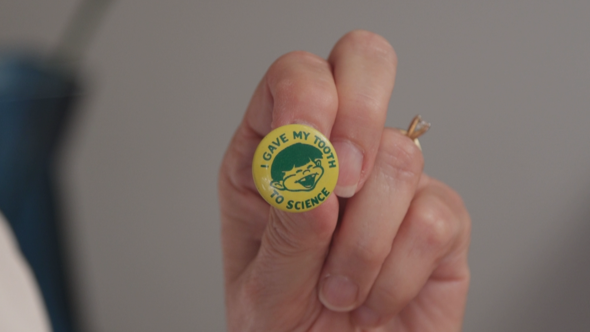 A hand holding a yellow badge with green text and a cartoon face. The badge says, 'I gave my tooth to science.'