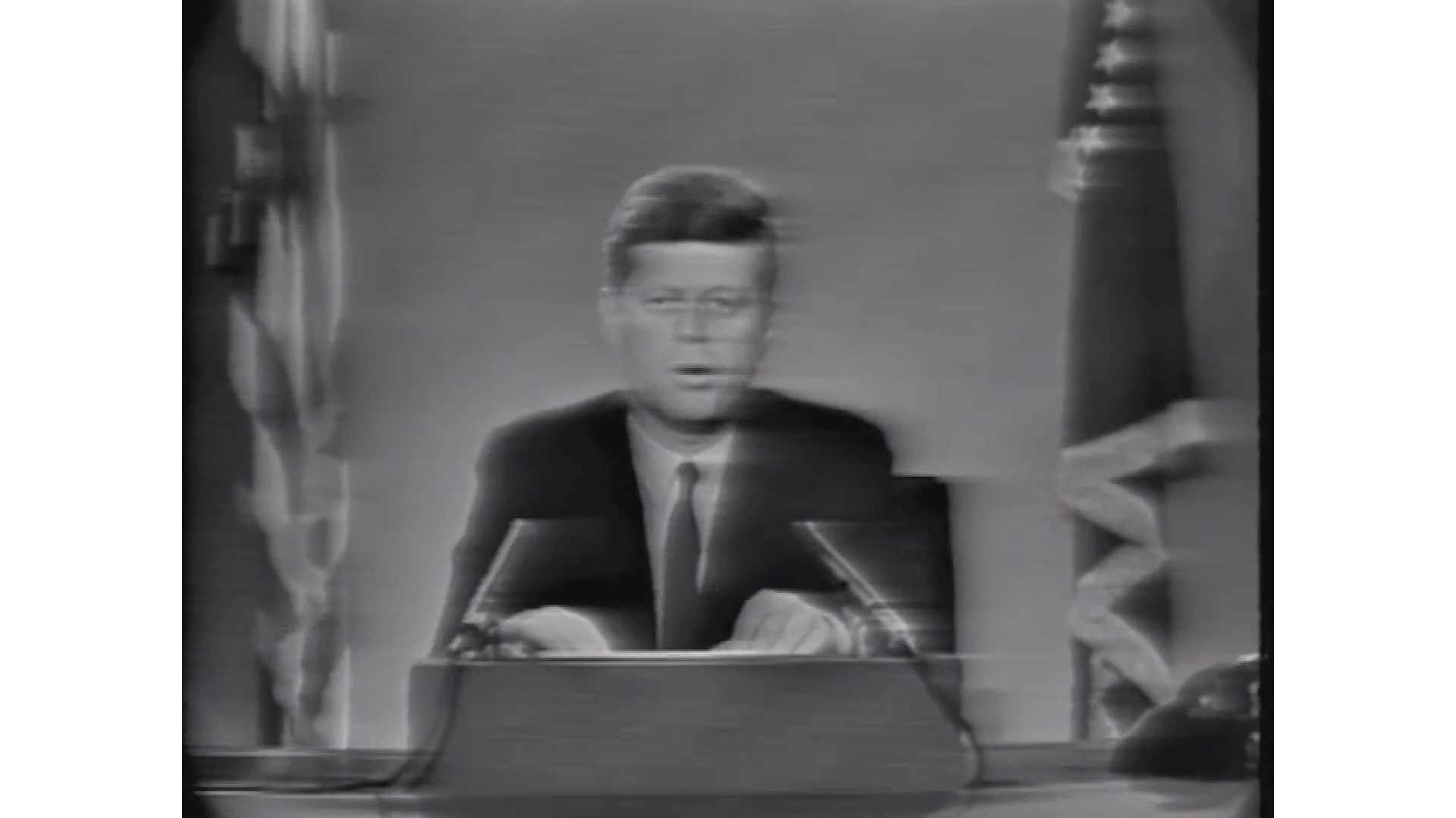 Black and white image of a man with glasses sitting at a desk, with flags partially visible on either side, appearing to be in a formal or news studio setting.