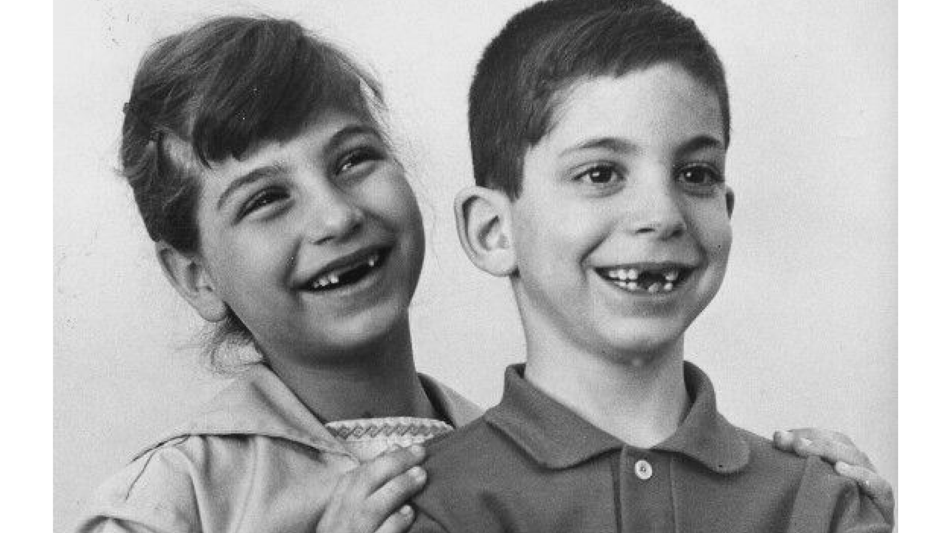 Two children, a girl and a boy, smiling and showing missing teeth, with the girl placing her hand on the boy's shoulder.