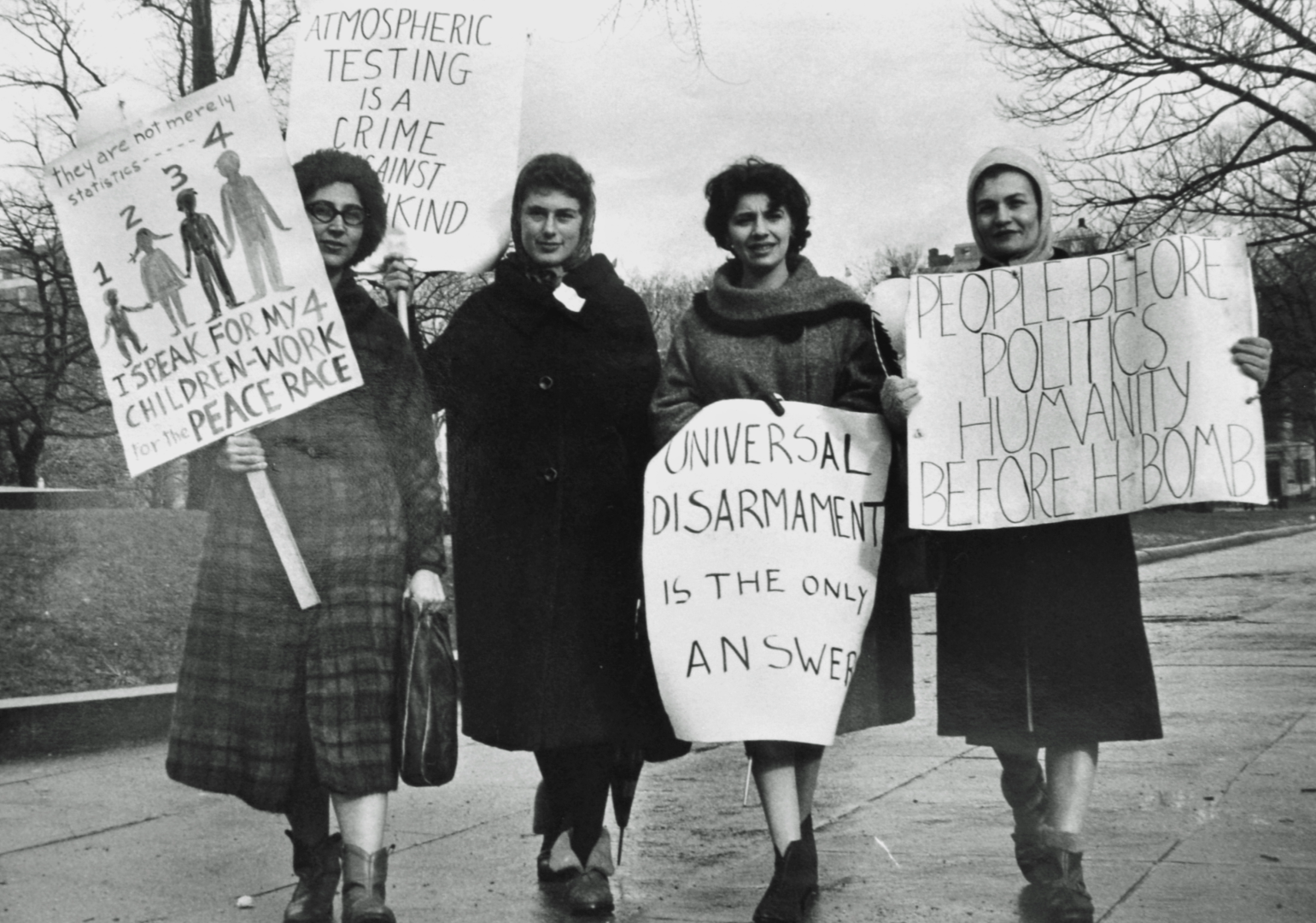 Four women protesting outdoors holding signs advocating for peace, disarmament, and anti-war messages, with leafless trees in the background.