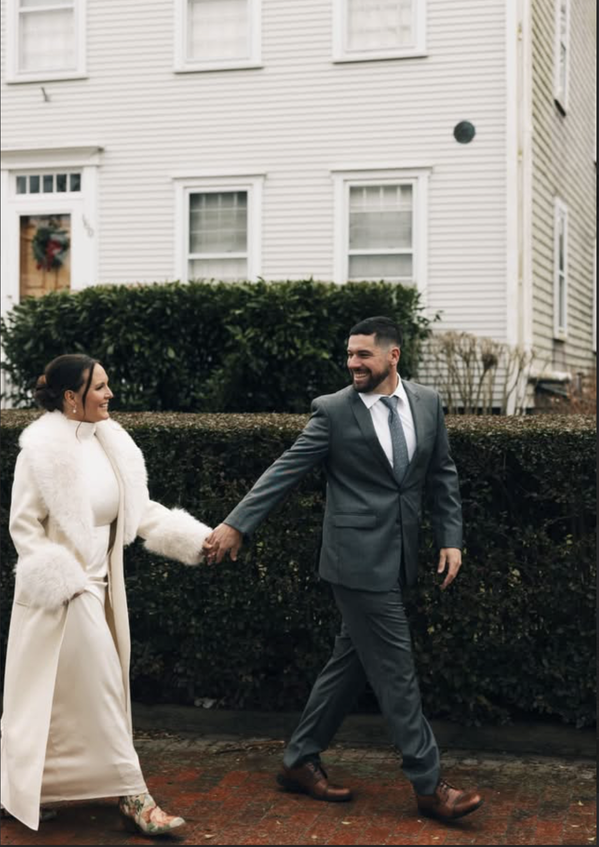 Couple holding hands on a sidewalk in front of a white house with hedges, dressed in formal winter attire, smiling and looking at each other.